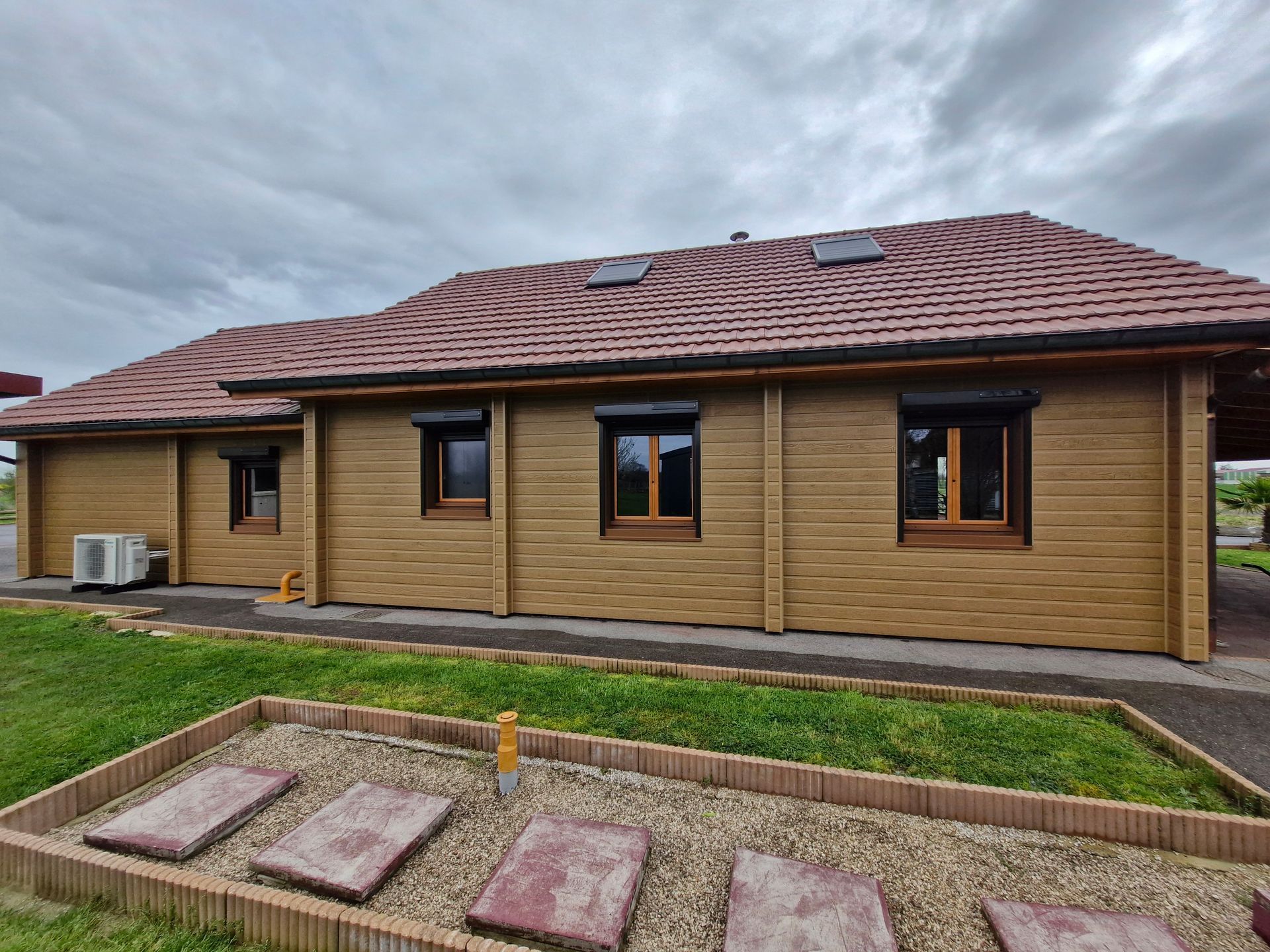 Bâtiment en bois avec toit en tuiles rouges, fenêtres sombres et élément de climatisation. Journée nuageuse.