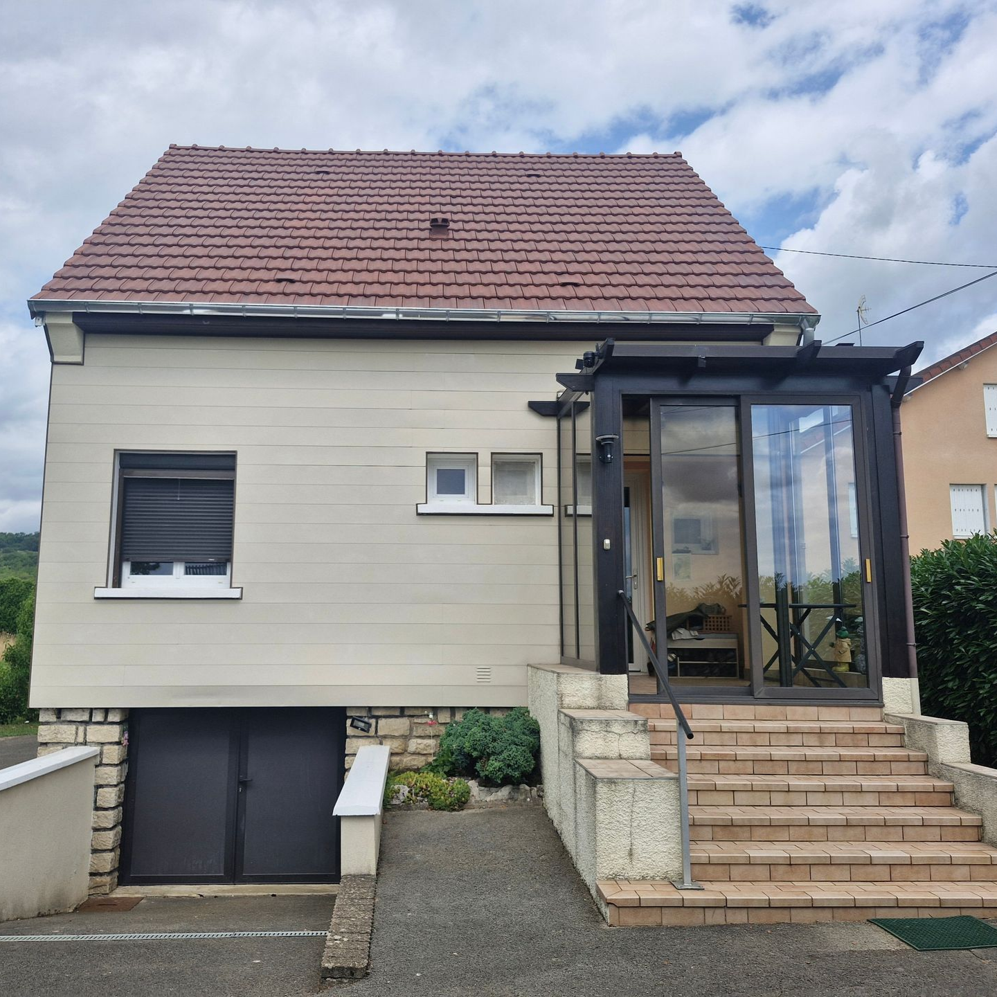 Maison aux façades beiges avec un toit rouge, une entrée vitrée, des marches et une porte de garage.