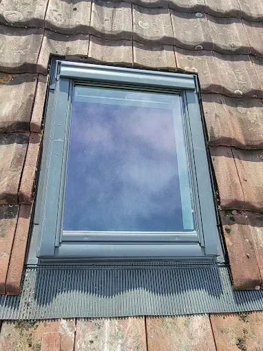 Puits de lumière dans un toit en tuiles de terre cuite, avec volet roulant fermé. Ciel bleu visible.