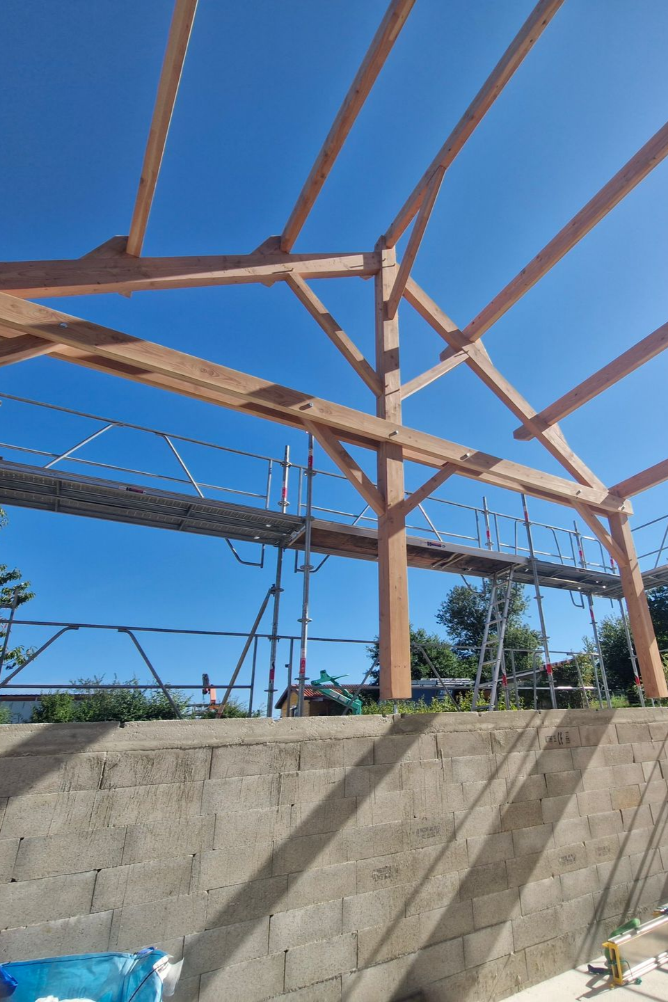 Ossature en bois d'un bâtiment en construction, sur fond de ciel bleu, soutenue par un mur de briques et un échafaudage.