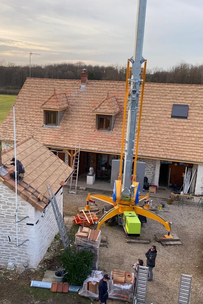 Une grue soulève des tuiles sur une maison en construction ; des ouvriers, des briques et des machines sont visibles.