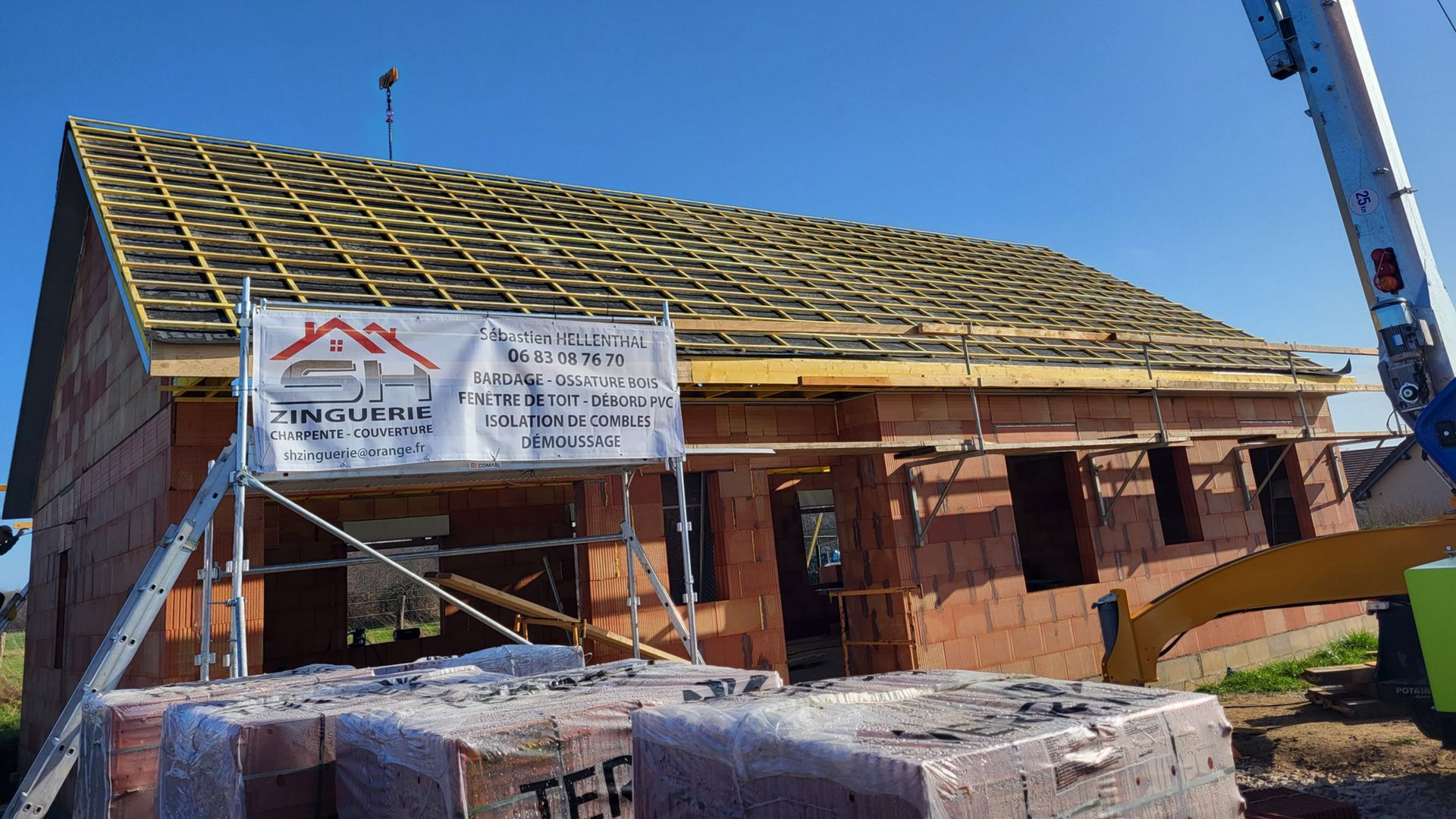 Maison en construction avec une charpente, des briques, un échafaudage et une pancarte devant un ciel bleu clair.