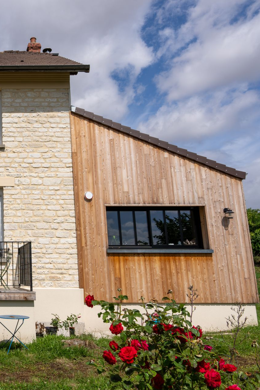 Vue latérale d'une maison avec une extension en bois, des rosiers devant, un ciel bleu au-dessus.