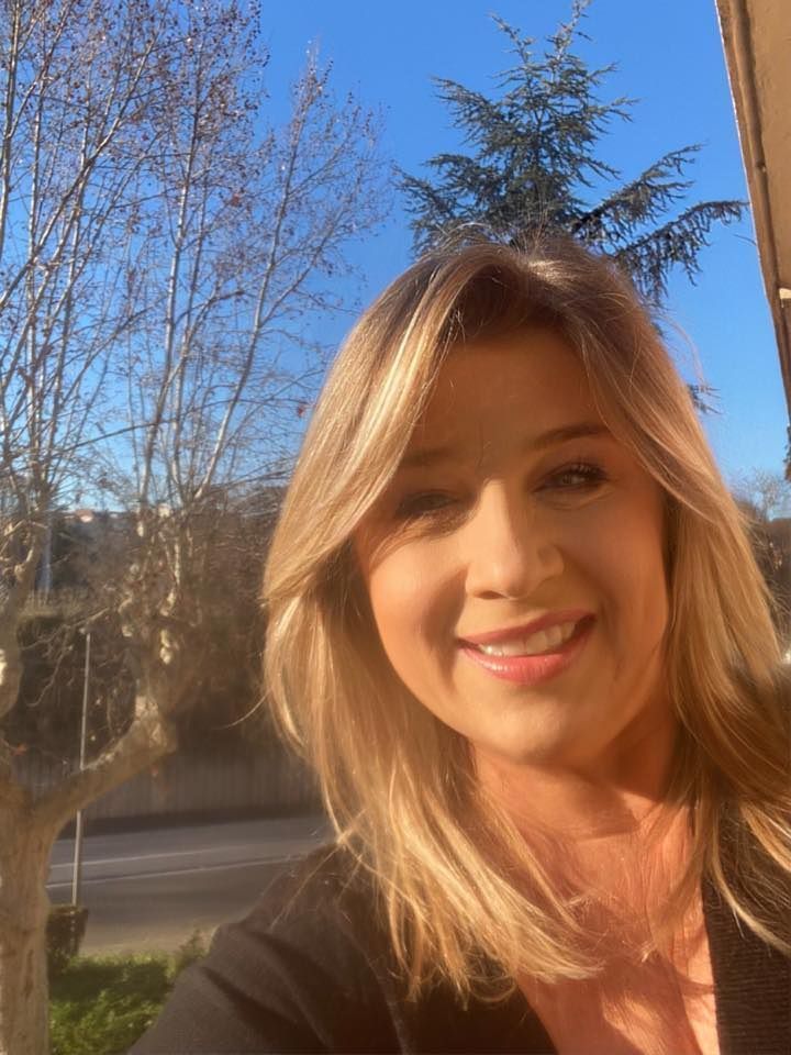 Une personne souriante aux cheveux blonds mi-longs, en extérieur.