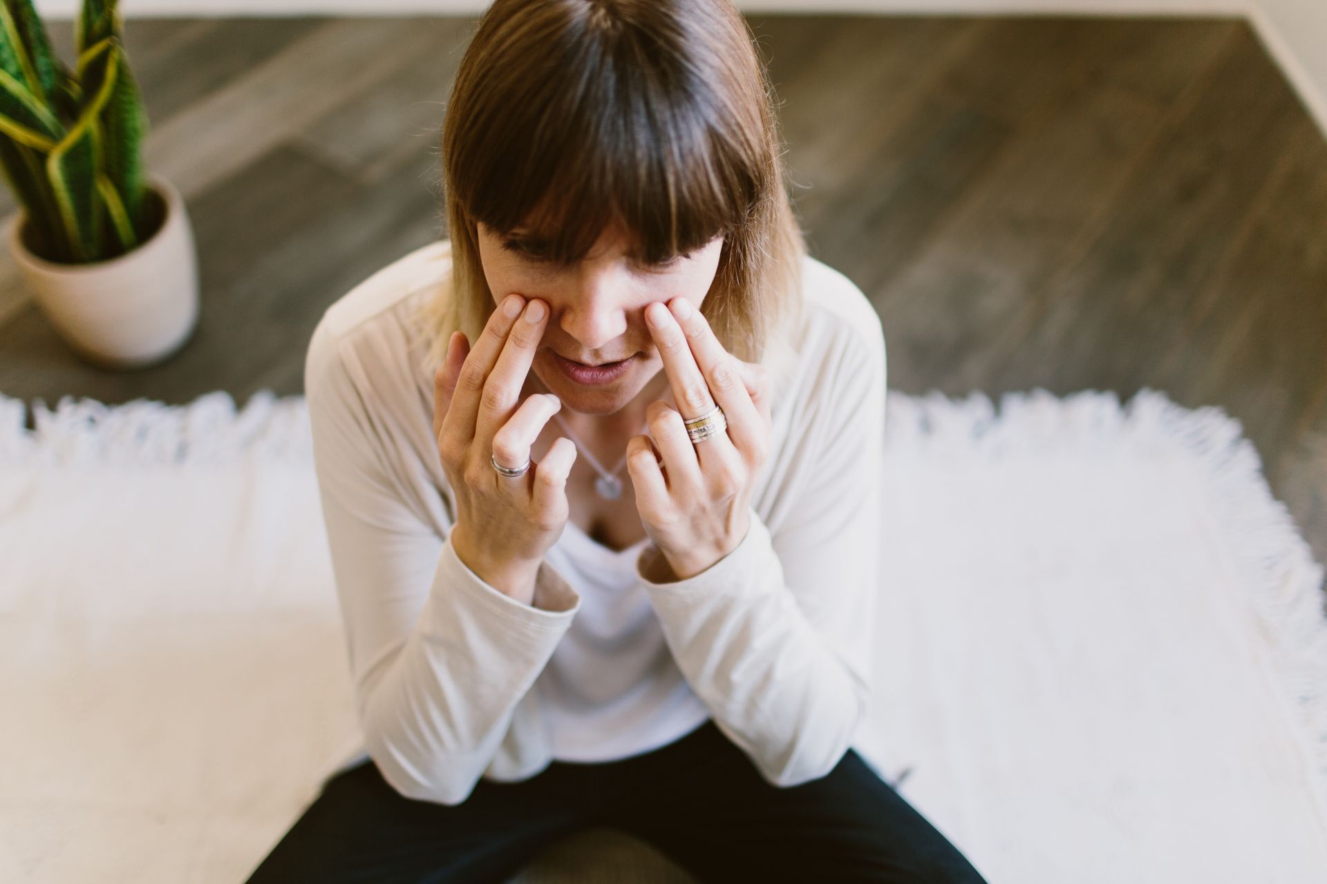 Une personne vêtue d'un haut crème, assise sur un tapis blanc, se touche le visage près du nez et des yeux, avec une plante en arrière-plan.