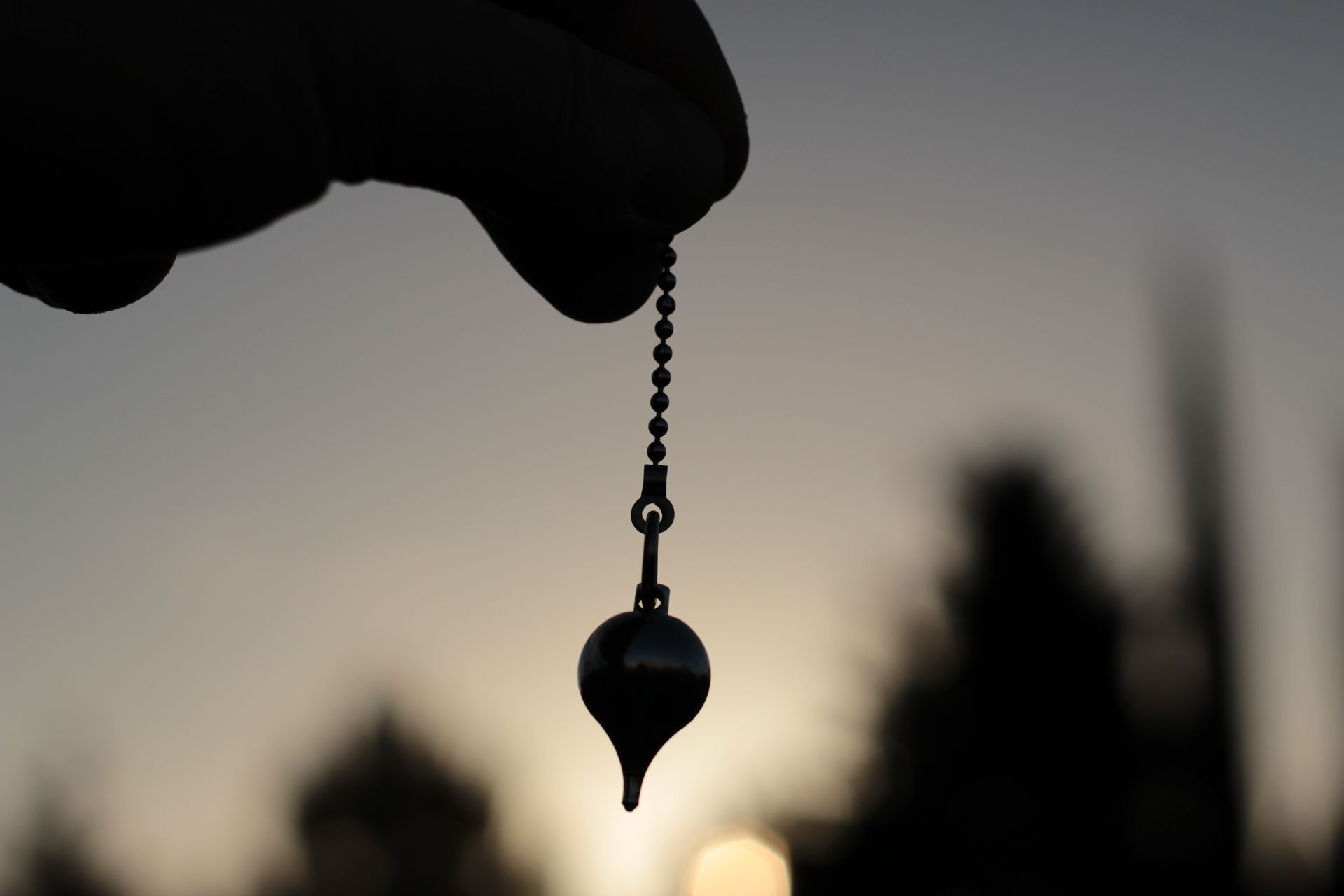 Une main tient un pendule métallique au bout d'une chaînette de perles, sur un fond de coucher de soleil.