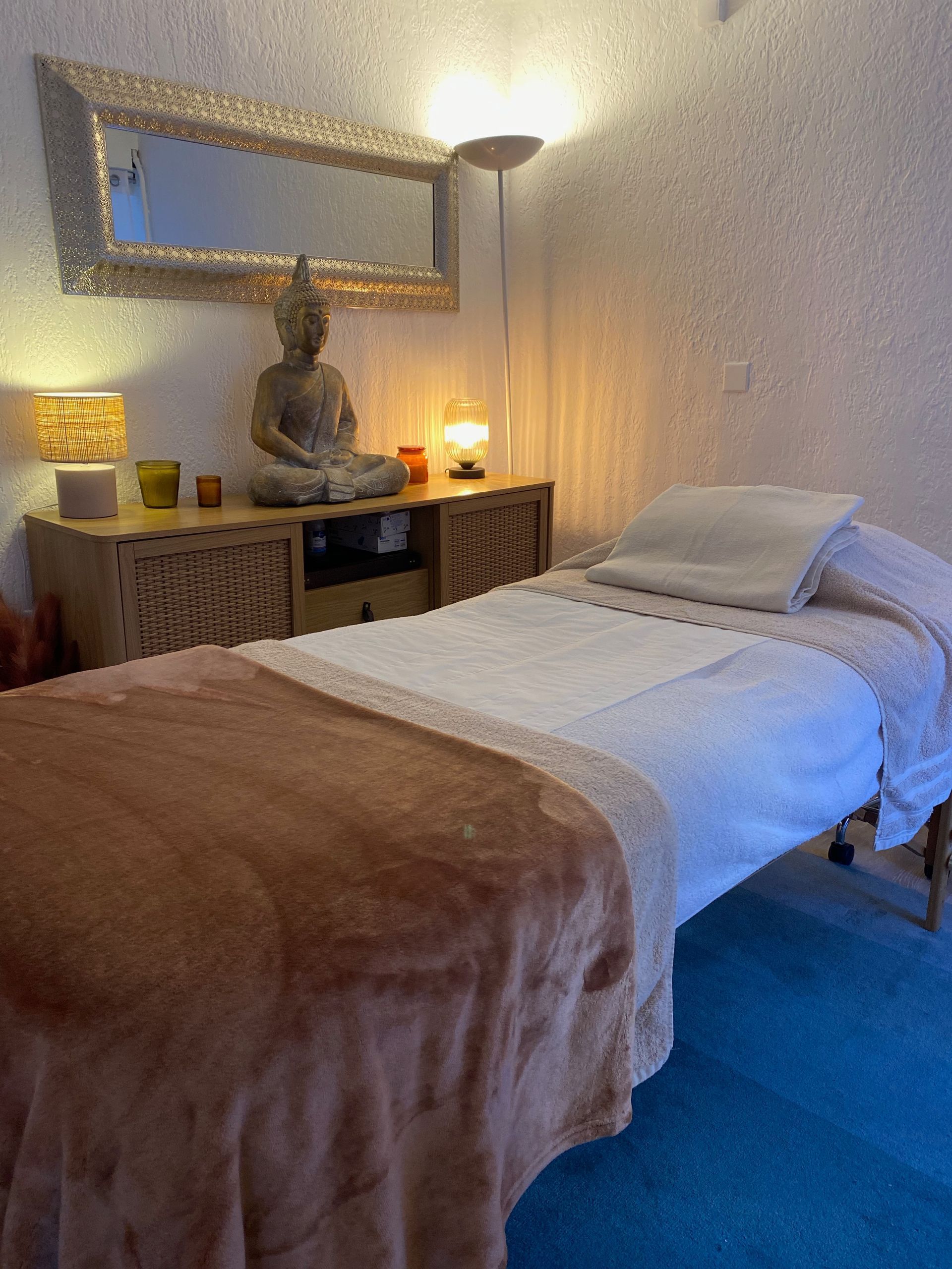 Une salle de soins avec une table de massage, une statue de Bouddha sur une armoire en bois, un éclairage tamisé et un miroir mural.