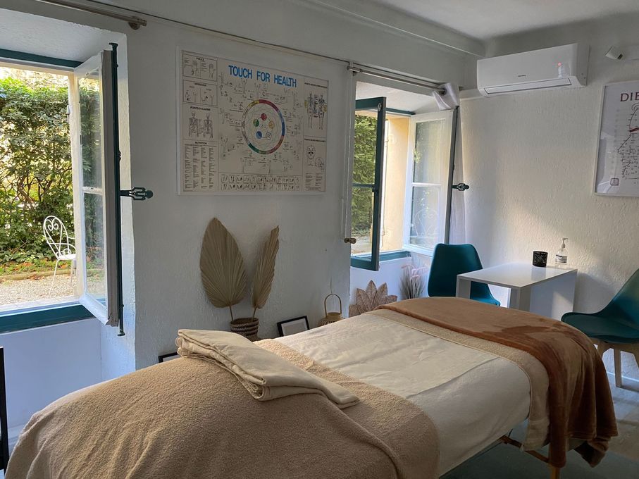 Une salle de massage avec une table de soins, du linge de lit texturé beige, des fenêtres ouvertes, des œuvres d'art murales et des chaises vertes.