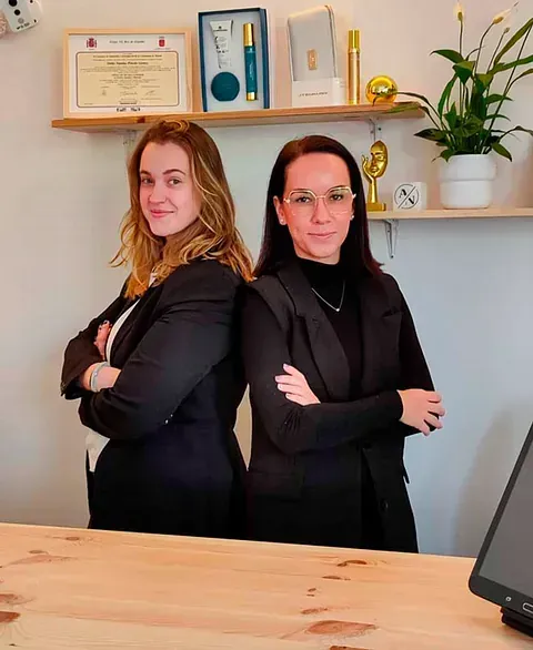 Dos mujeres están de pie, espalda con espalda, frente a un escritorio, con los brazos cruzados.