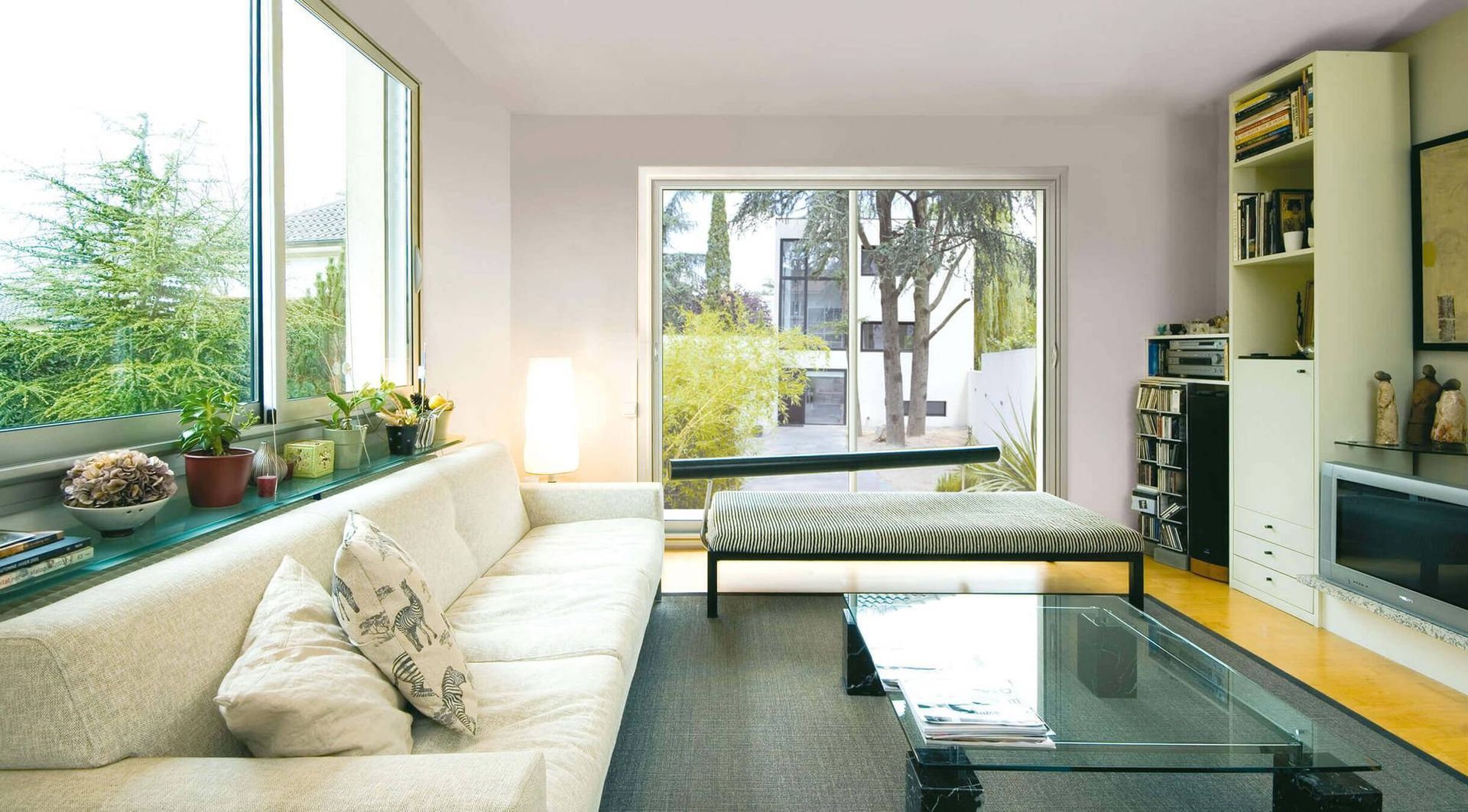 Salon moderne et lumineux avec canapé blanc, table basse en verre, télévision murale et grandes fenêtres donnant sur un jardin.