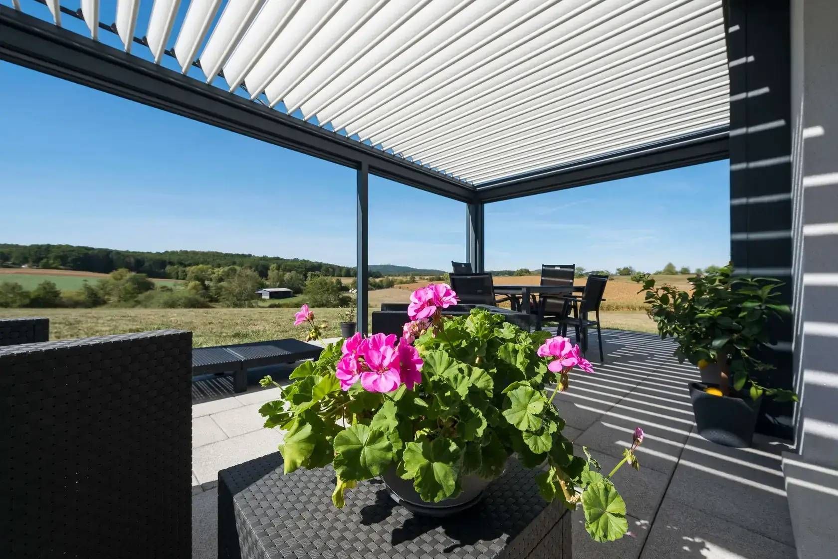 Patio avec chaises en osier noir, fleurs roses et vue sur la campagne sous une pergola à claire-voie
