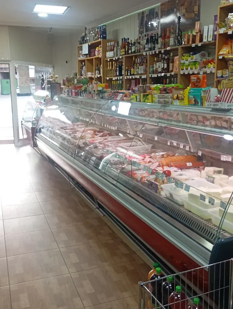 Dentro de una tienda de delicatessen, vitrina repleta de carnes y quesos, estantes de productos al fondo.