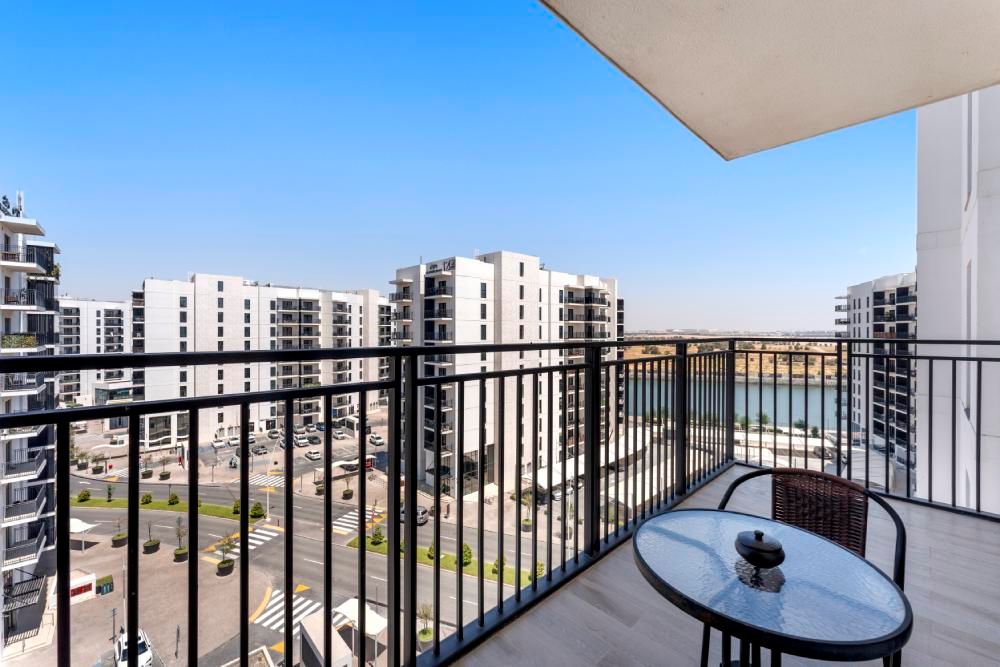 Balcony with glass table and chairs overlooking modern apartment buildings and a pool under a blue sky
