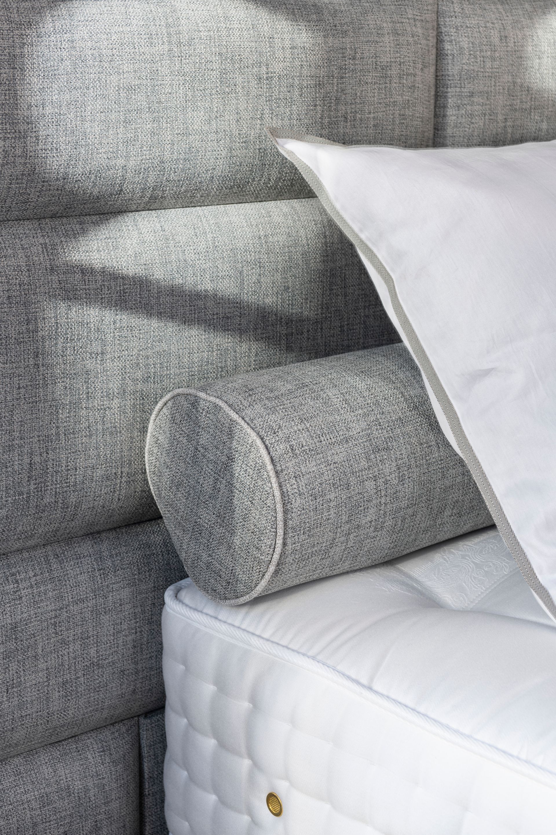 Nahaufnahme eines Bettes mit grauem, gepolstertem Kopfteil, Nackenrolle und weißer Bettwäsche. Nahaufnahme eines Bettes mit grauem, gepolstertem Kopfteil, Nackenrolle und weißer Bettwäsche.