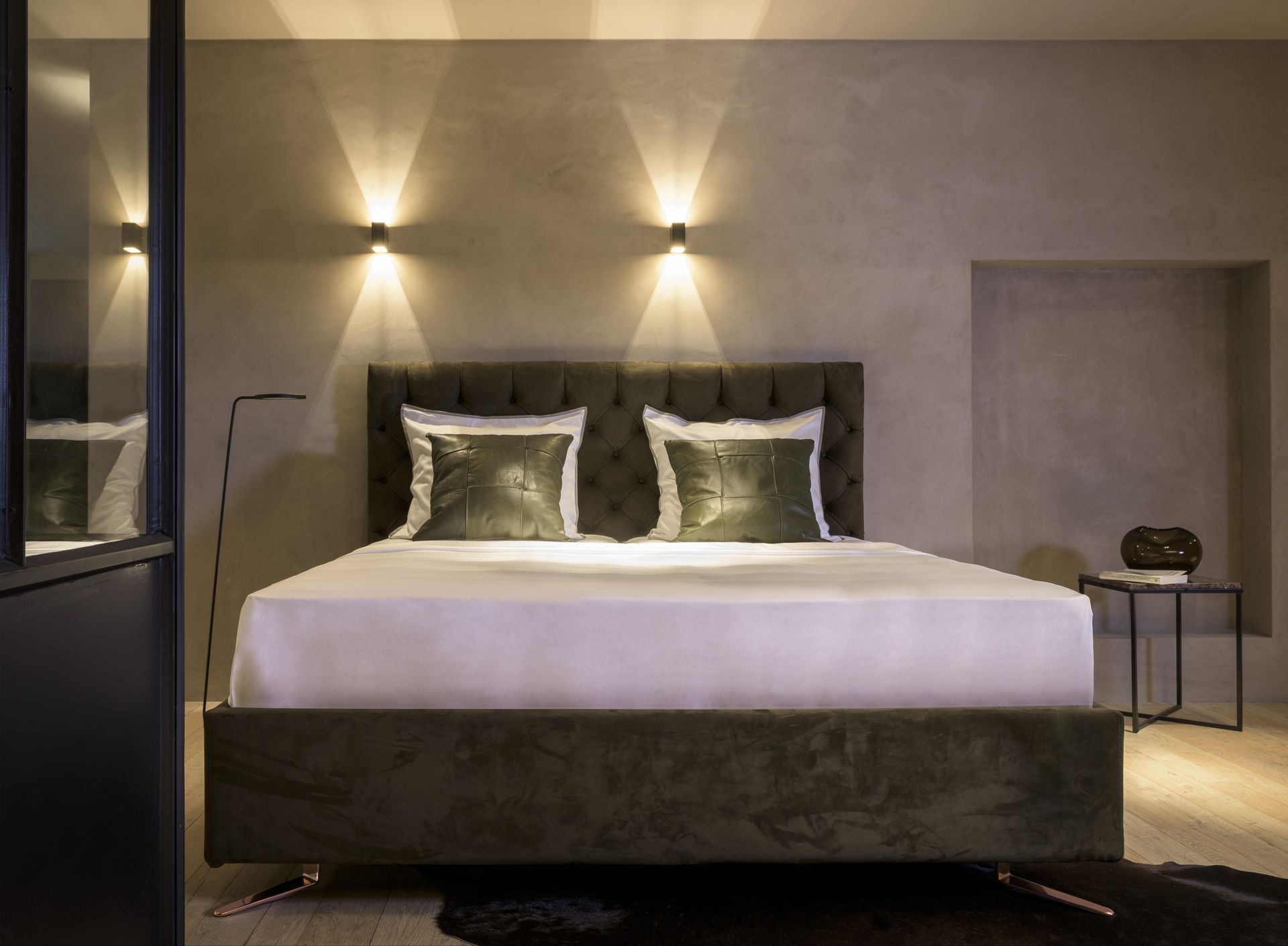 Dunkelgrünes Samtbett mit weißen Laken und Kissen, beleuchtet von Wandleuchten in einem Schlafzimmer.