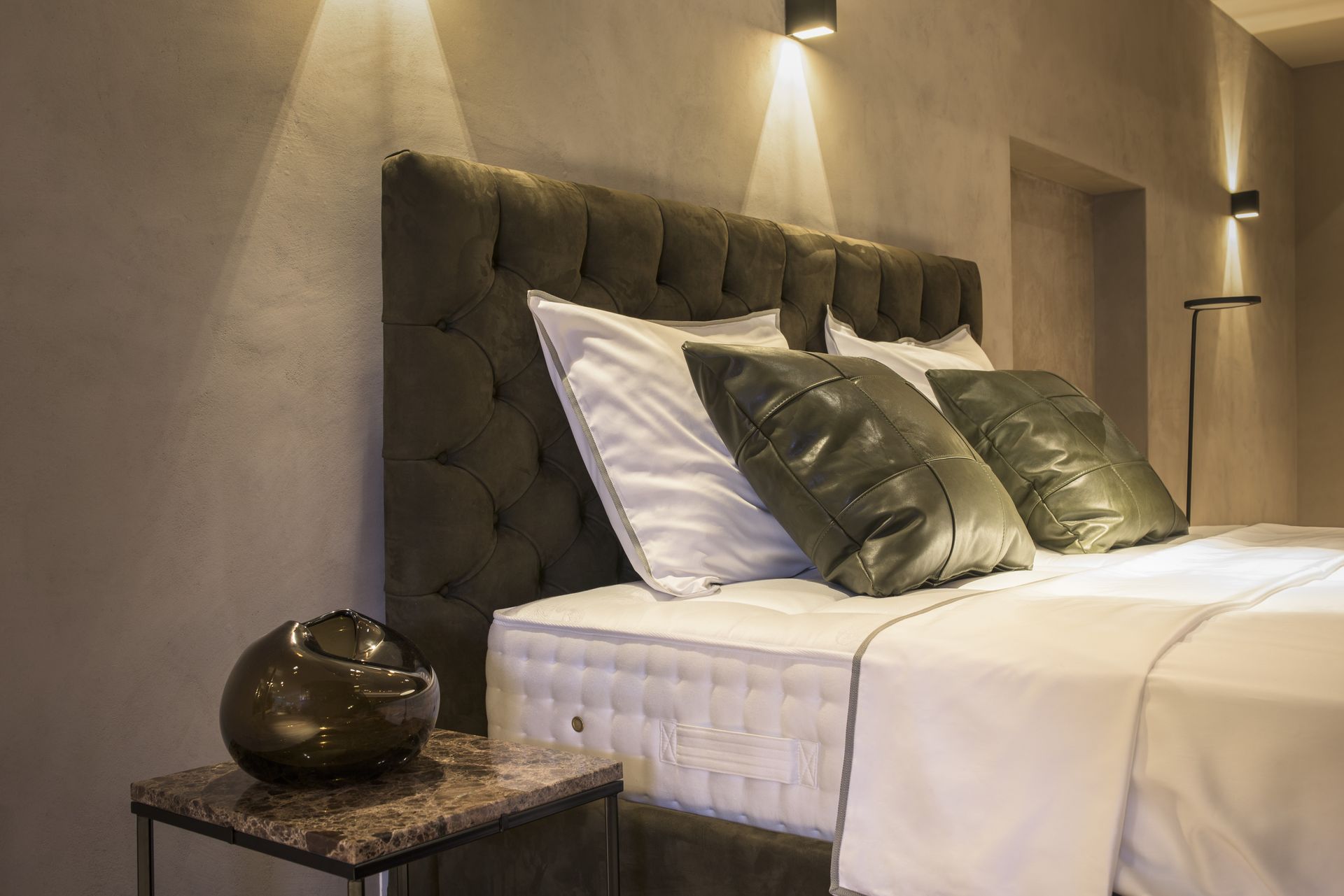 Schlafzimmer mit gestepptem Kopfteil, strahlend weißer Bettwäsche, grünen Kissen, Beistelltisch mit dekorativer Vase und Wandleuchten.