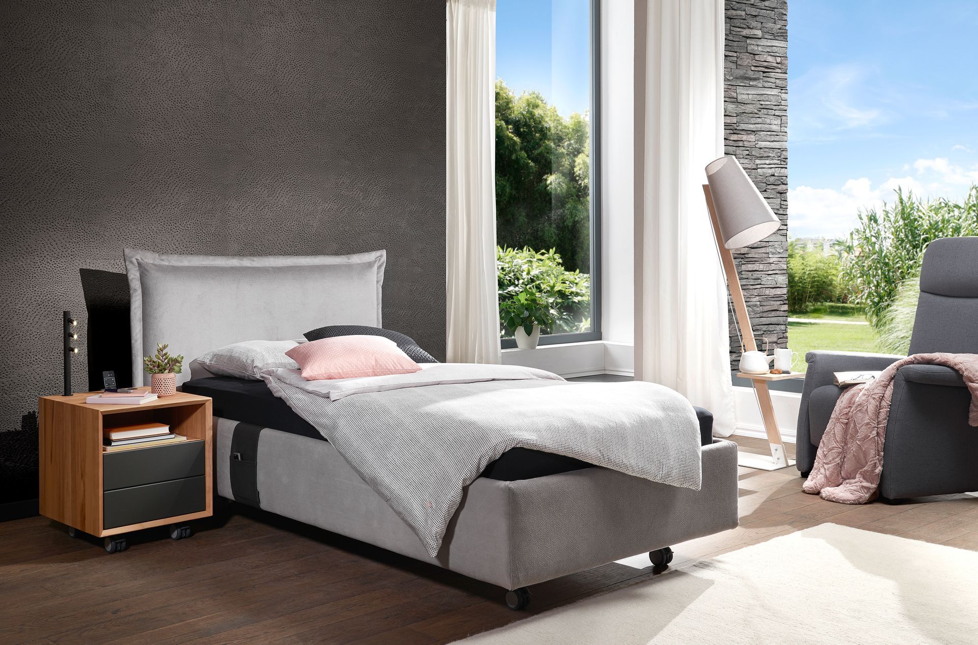 Schlafzimmer mit grauem Bett, Nachttisch, Sessel und Fenster mit Blick auf einen Garten.