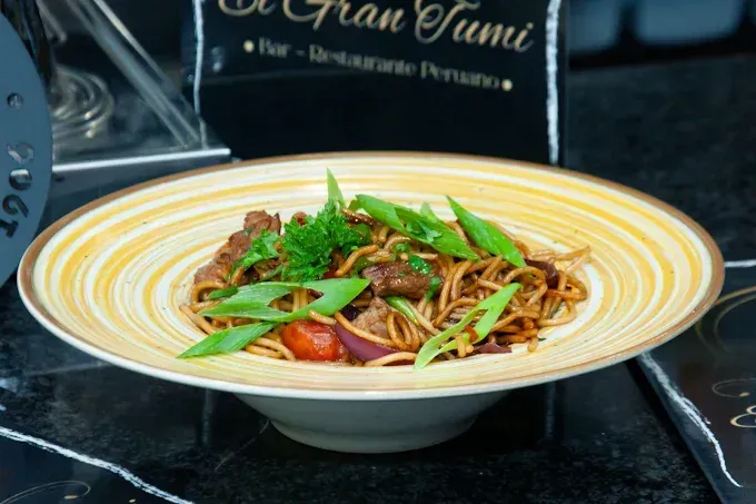 Plato de fideos salteados con carne, verduras y cebollino. Ambiente de restaurante, tonos marrones y beige.