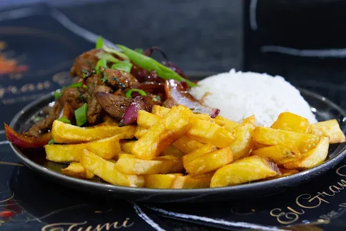 Plato de comida: carne de res salteada con cebolla roja y cebolletas, servida con arroz blanco y papas fritas.