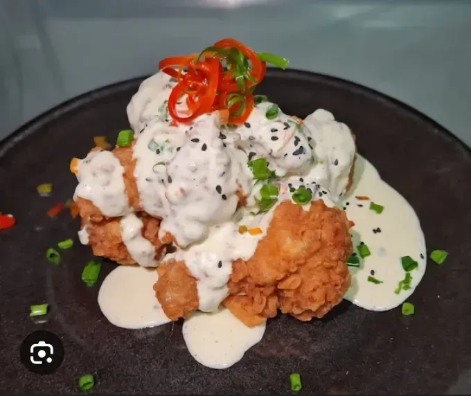 Coliflor frita con salsa cremosa, adornada con cebolletas, pimientos rojos y semillas de sésamo negro en un plato negro.