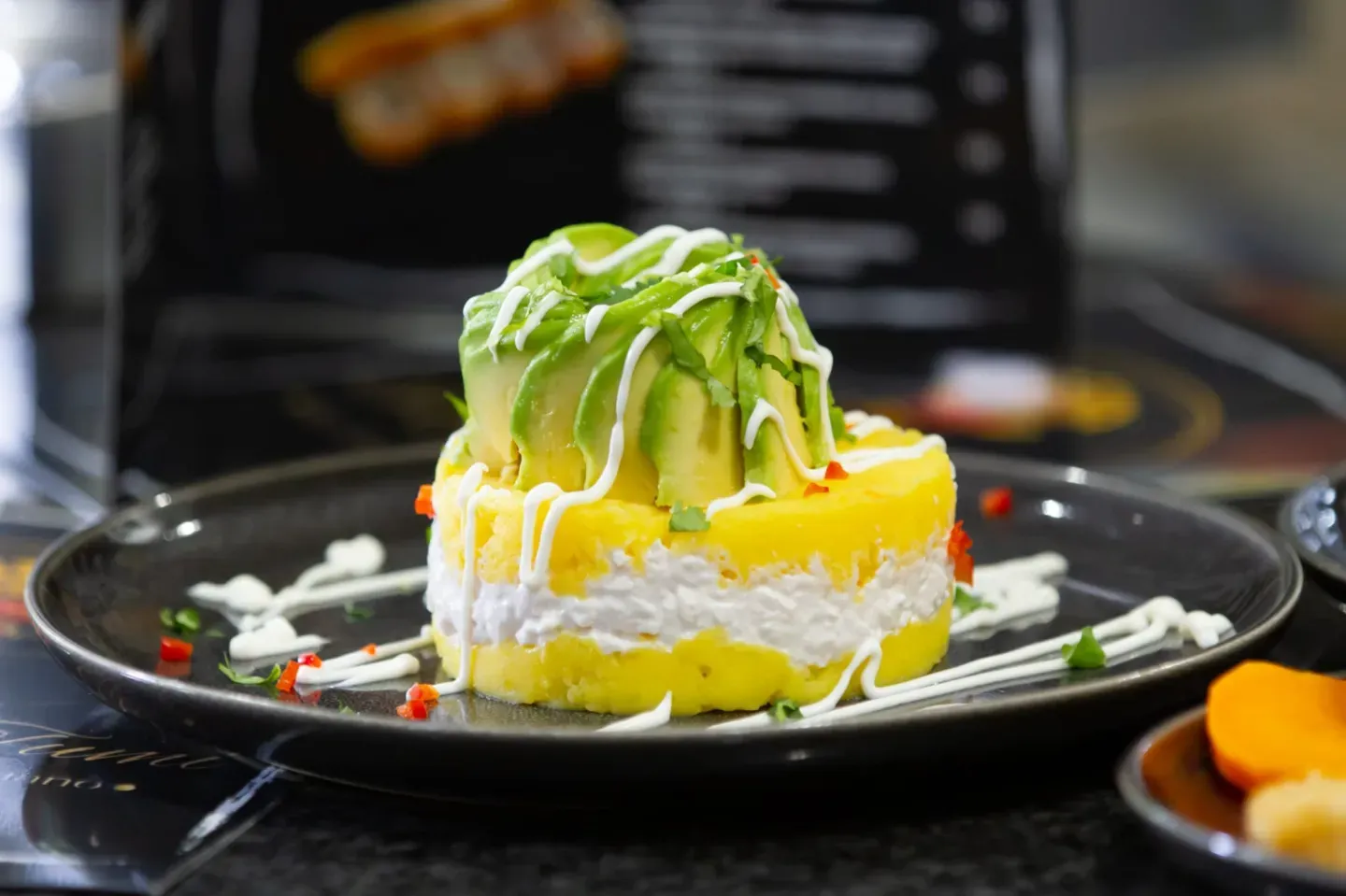 Plato de causa peruana en capas con aguacate, sobre plato negro, decorado con salsa blanca.