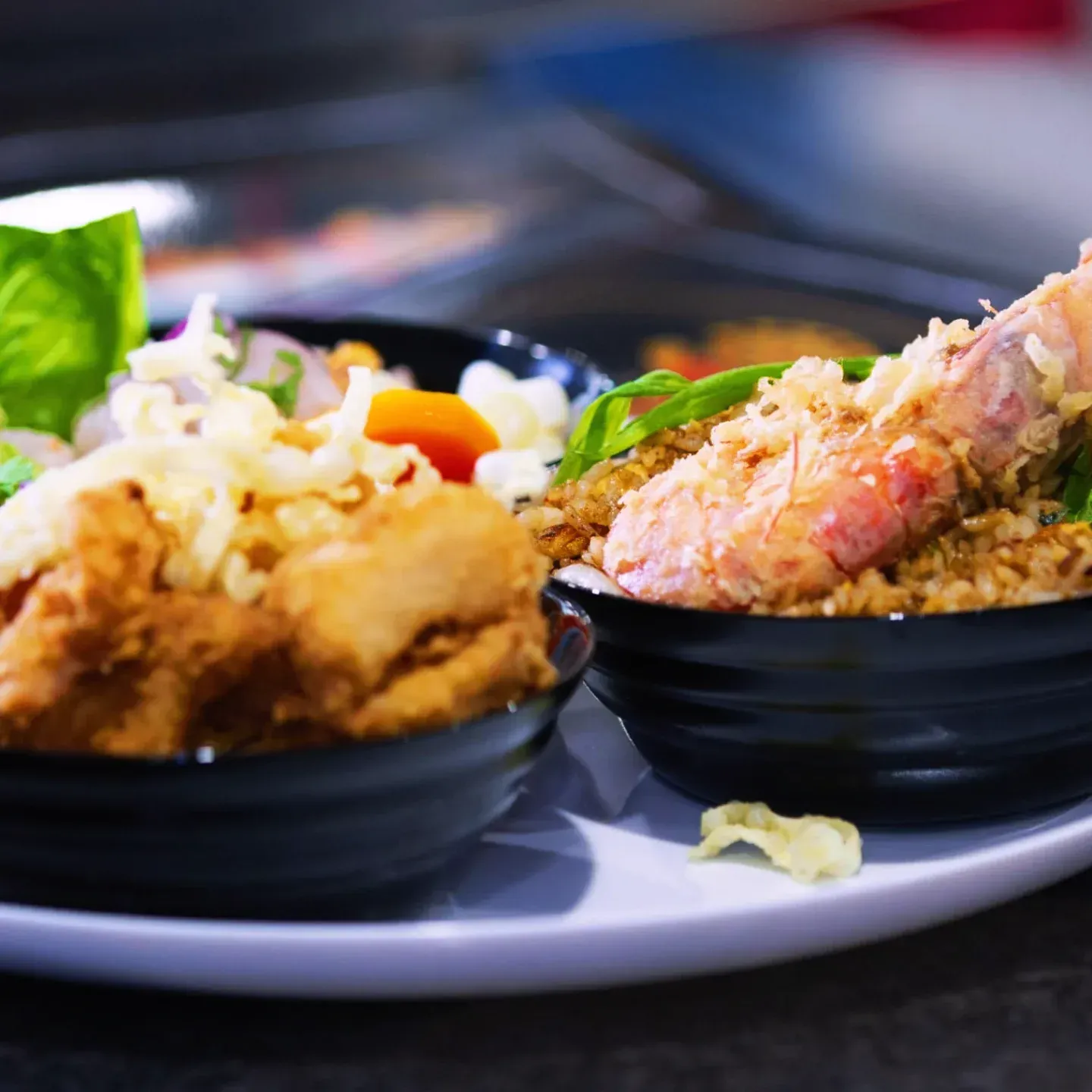 Dos tazones negros de comida en un plato blanco. Uno contiene pollo frito y el otro, camarones fritos.