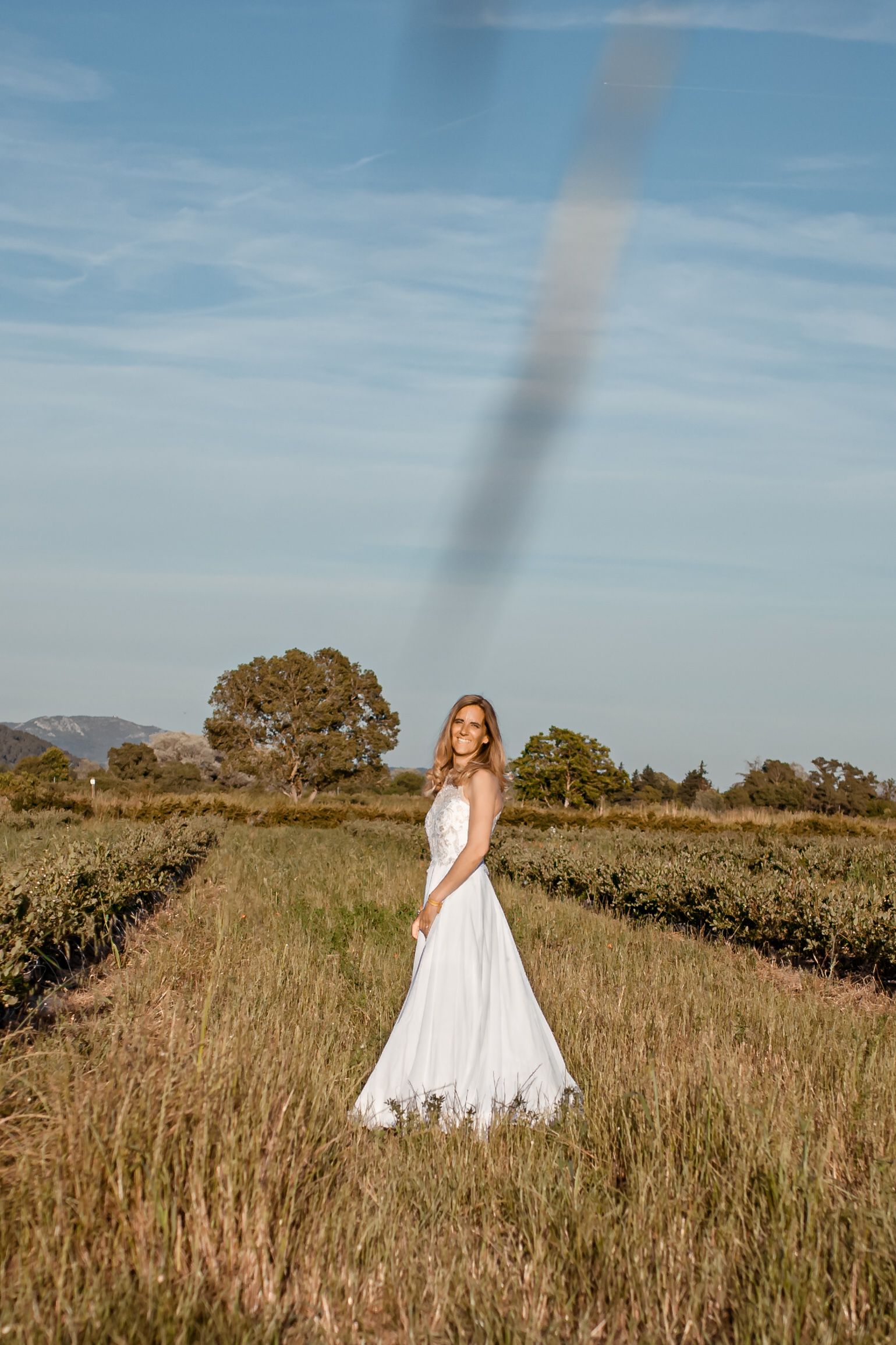 Une femme en robe blanche se tient dans un champ sous un ciel bleu ; des rangées de plantes sont visibles.