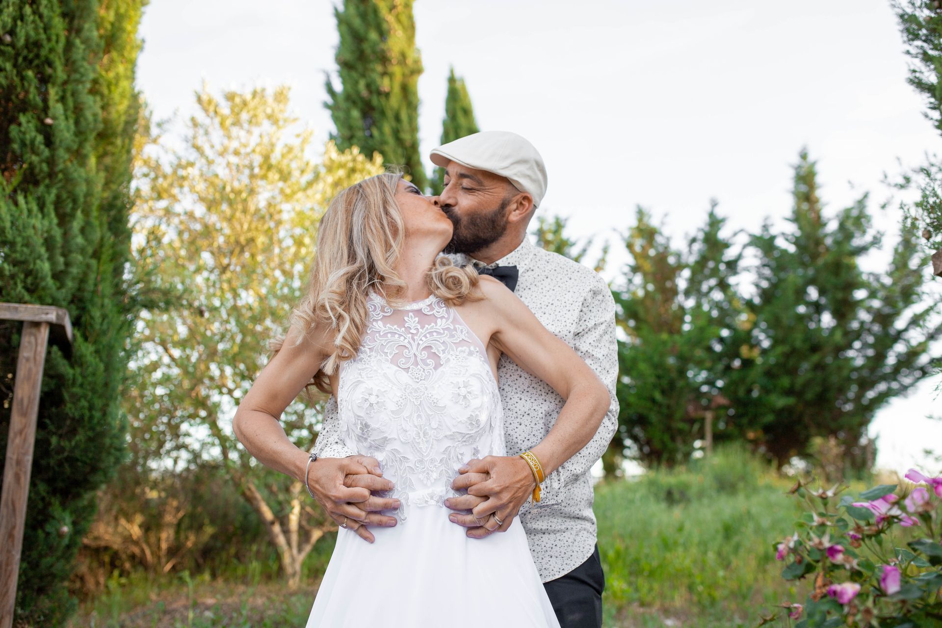 Couple s'embrassant en plein air ; la mariée en robe blanche, le marié en chapeau et nœud papillon, entouré de verdure.