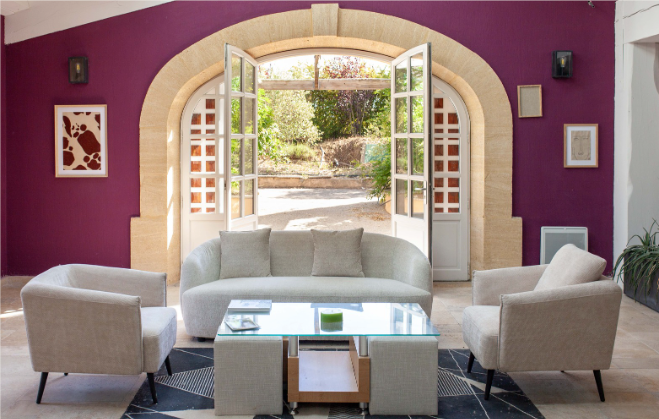 Salon avec mobilier couleur crème, table basse en verre et porte cintrée donnant sur le jardin. Murs violets.