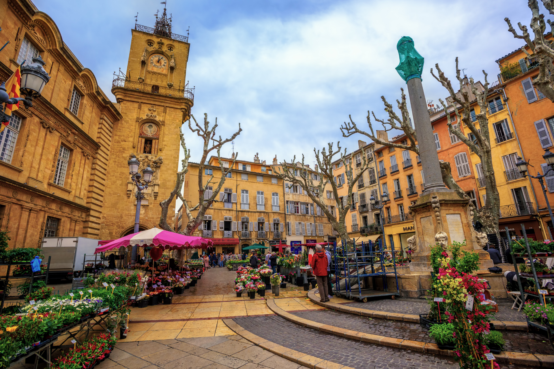 Un marché sur une place européenne avec une tour de l'horloge, des bâtiments, des gens et des étals de fleurs.