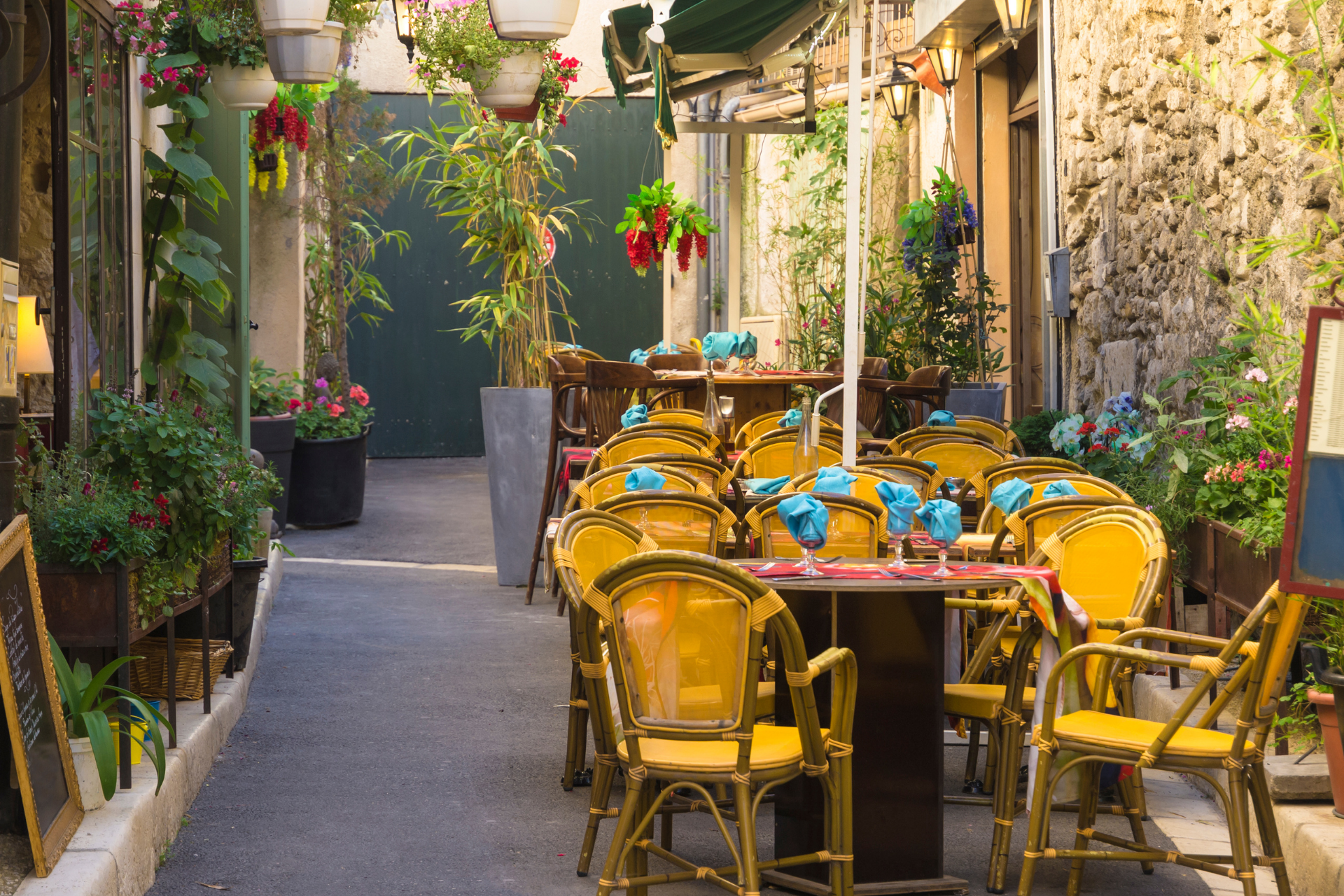 Ruelle étroite avec des sièges de café en plein air, des chaises jaunes, des plantes en pot et un mur de pierre.