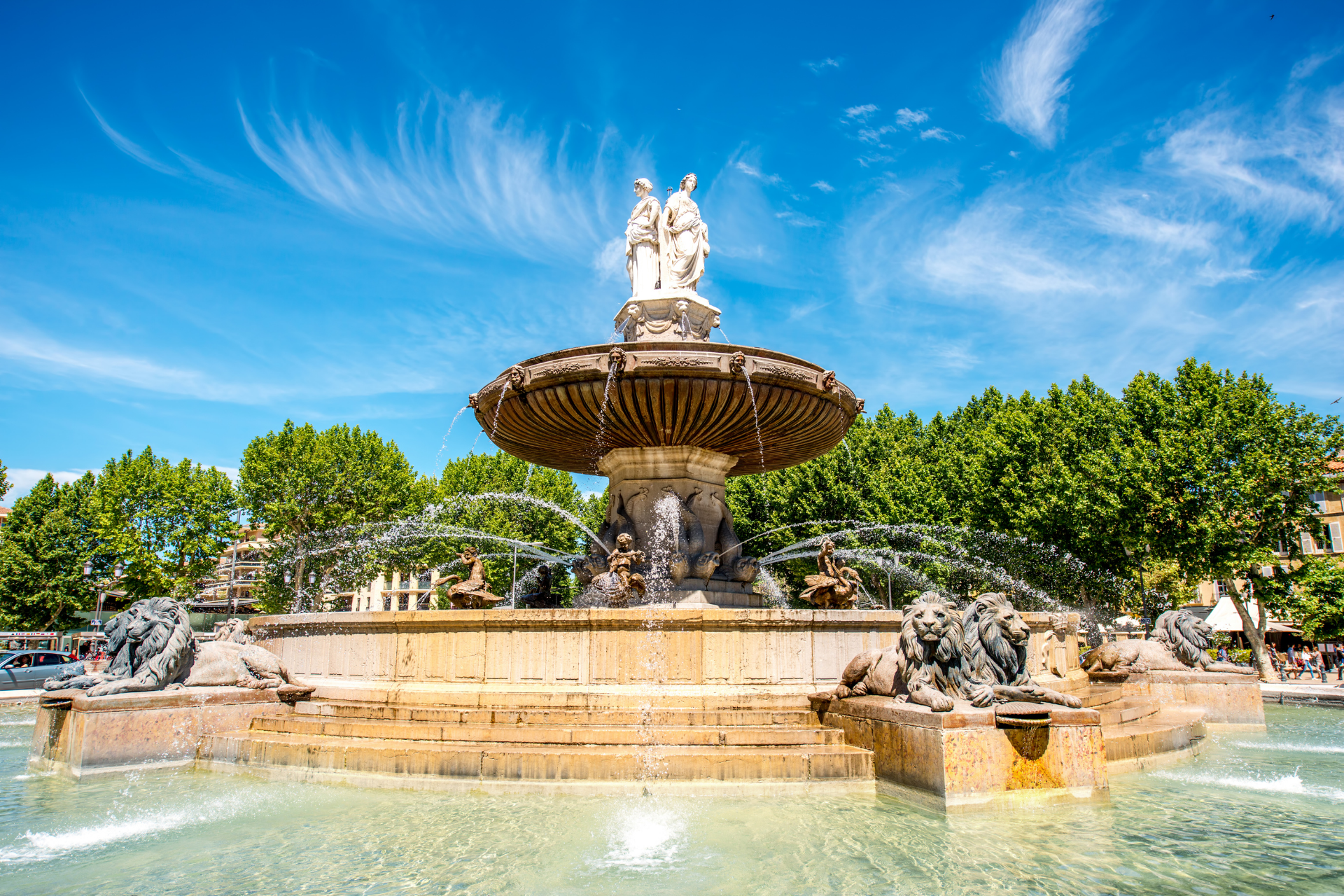 Fontaine sur une place ensoleillée avec une statue, de l'eau qui pulvérise et des arbres contre un ciel bleu vif.
