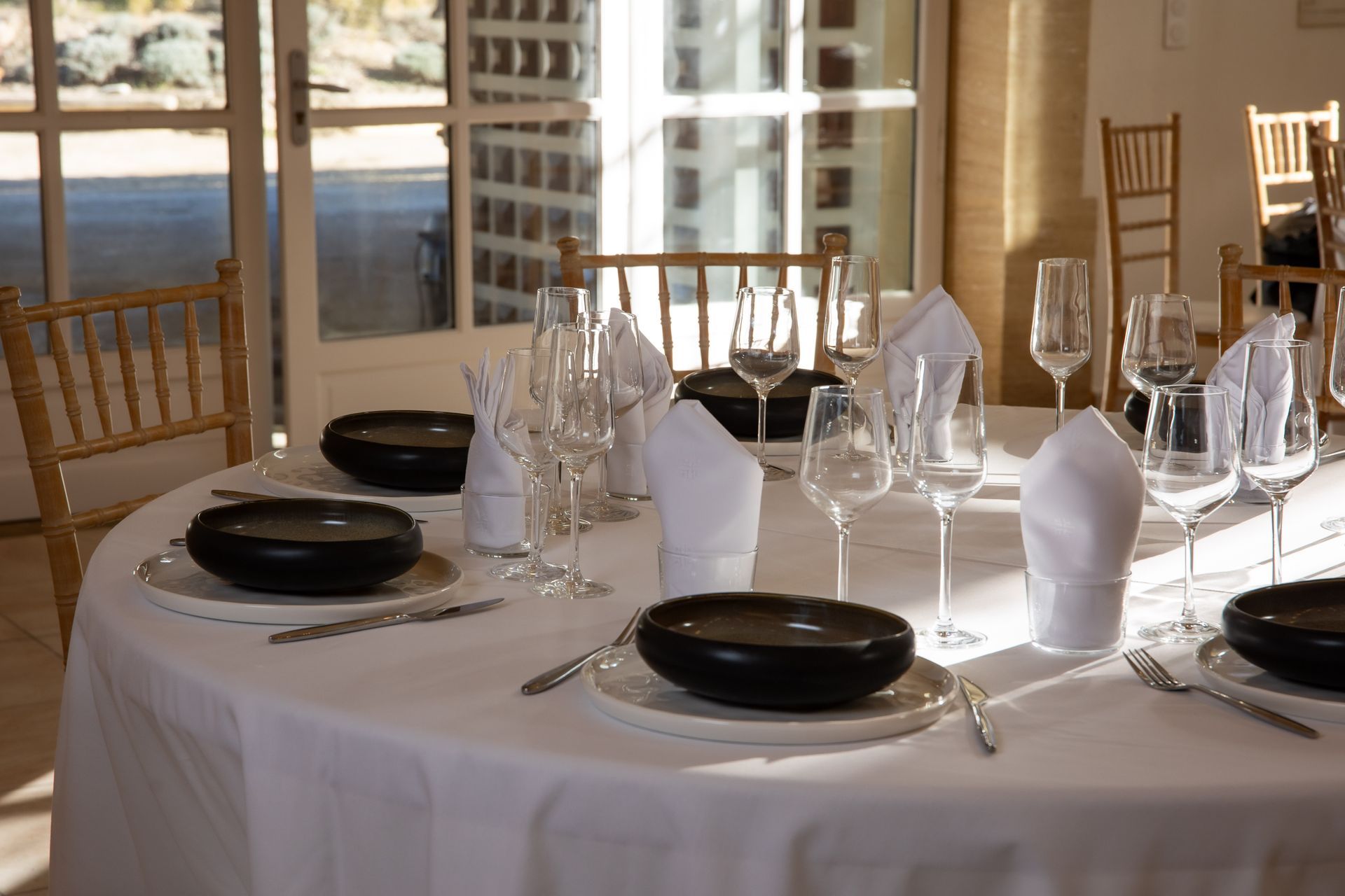 Table ronde dressée pour un dîner formel avec nappes blanches, assiettes, bols noirs et verres.