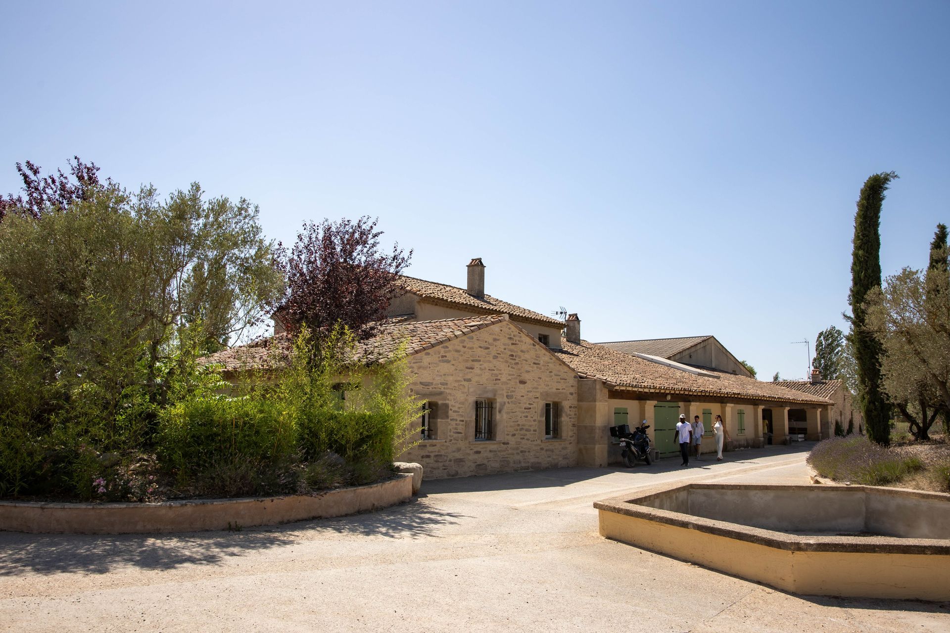 Bâtiment en pierre avec toit en tuiles, entouré d'arbres et d'une fontaine par une journée ensoleillée.