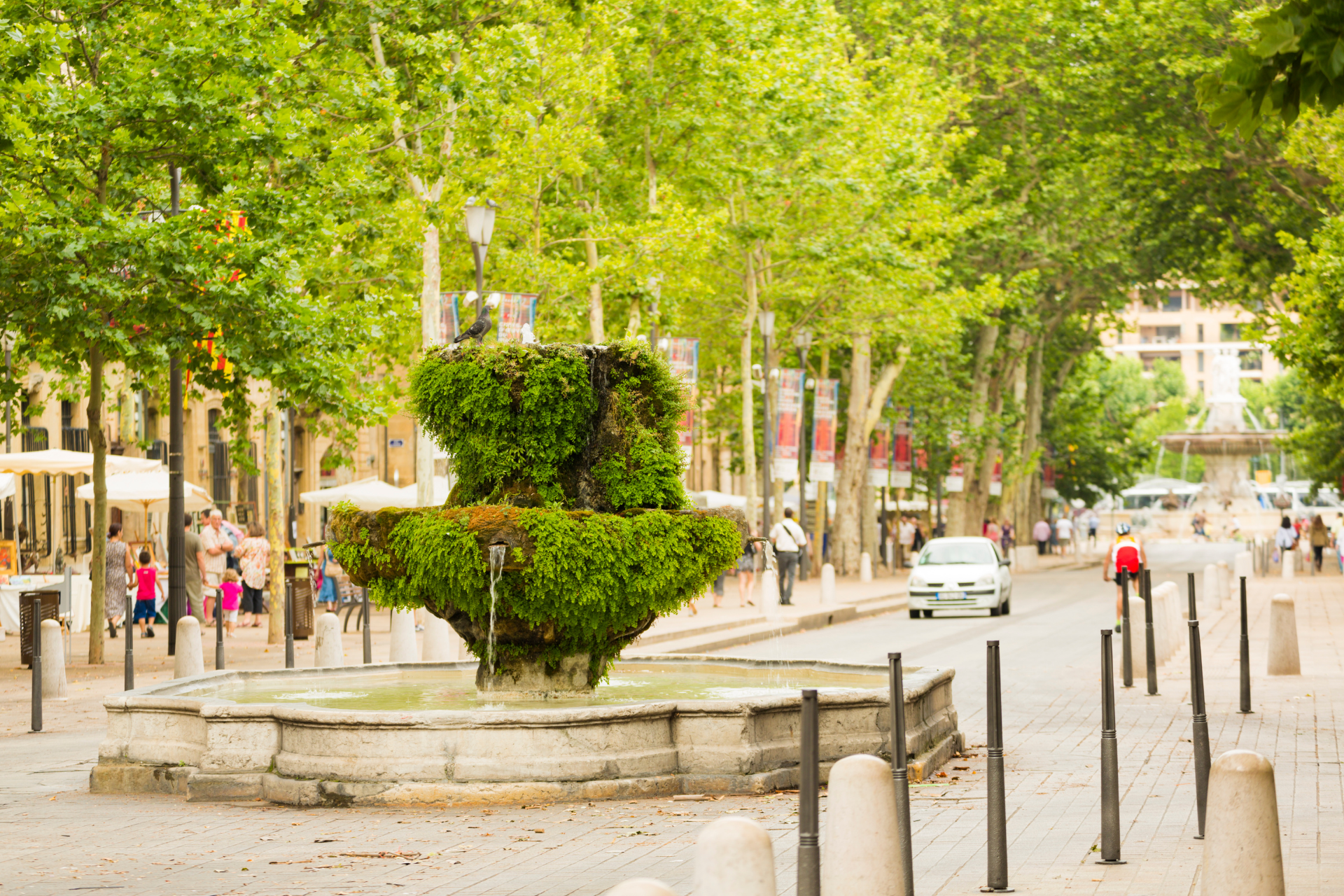 Une fontaine recouverte de lierre se trouve dans une rue de la ville bordée d'arbres et de piétons.
