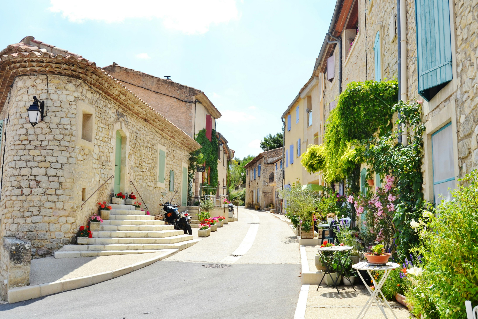 Des bâtiments en pierre bordent une rue étroite d'un village français, avec des fleurs et de la verdure.
