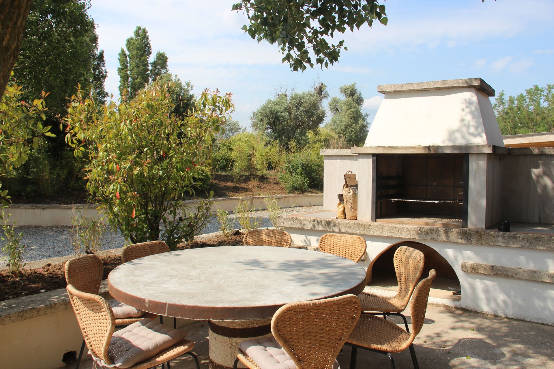 Terrasse extérieure avec une table en béton, des chaises tressées et un barbecue en pierre intégré sous un ciel bleu.