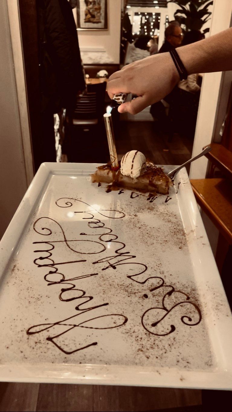 Person gießt Flüssigkeit über ein Dessert mit der Aufschrift „Happy Birthday“ in Schokolade auf einer Platte.