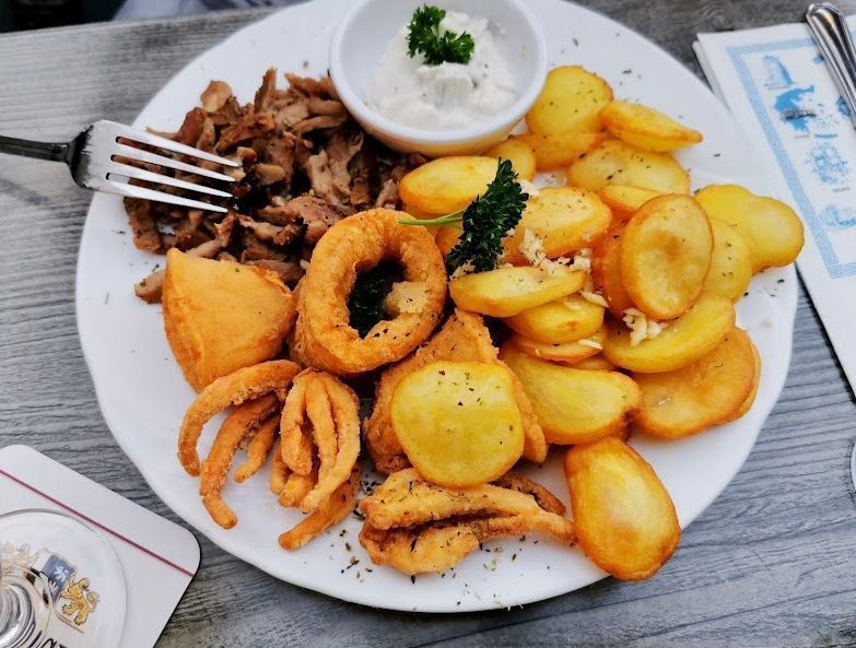 Teller mit Essen: Gyrosfleisch, Bratkartoffeln, Zwiebelringe, Soße und eine Gabel.