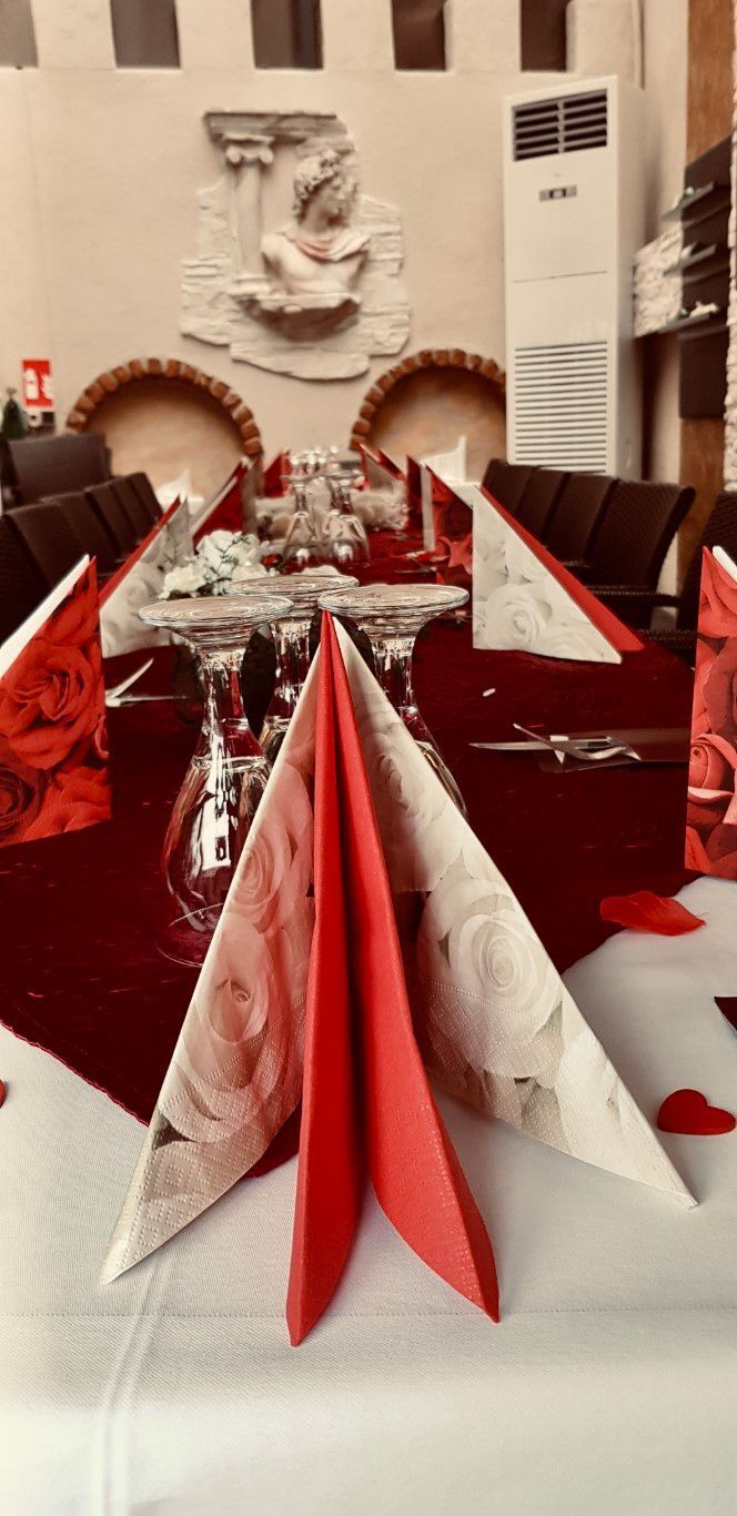 Restauranttisch mit roten und weißen Servietten, Blumen und Dekorationen gedeckt; im Hintergrund befindet sich eine Skulptur.