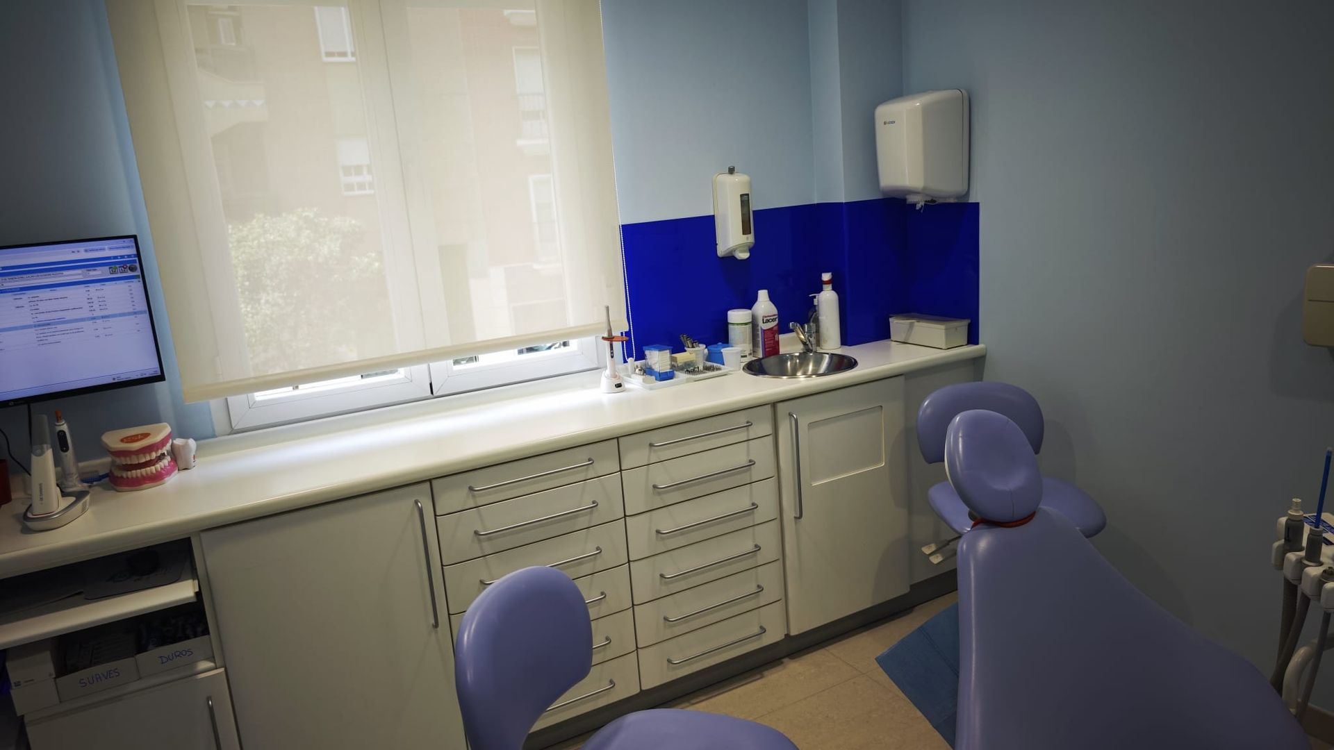 Consultorio dental con silla morada, mostrador, lavabo y equipo médico junto a una ventana.