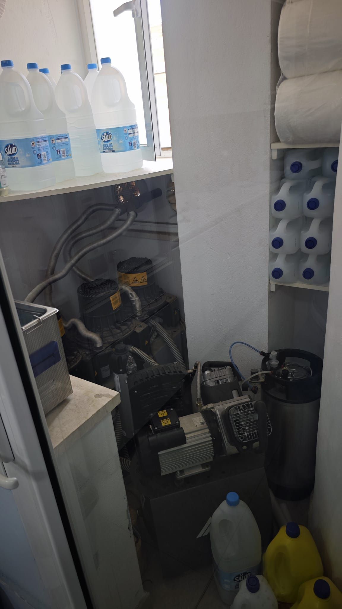 Cuarto de servicio desordenado con garrafas de agua, productos de limpieza, cables y equipos en estantes y en el suelo.