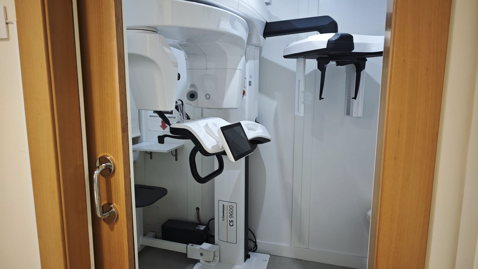 Máquina de rayos X dental en una pequeña sala de clínica con equipo blanco y gris, enmarcada por una puerta de madera.
