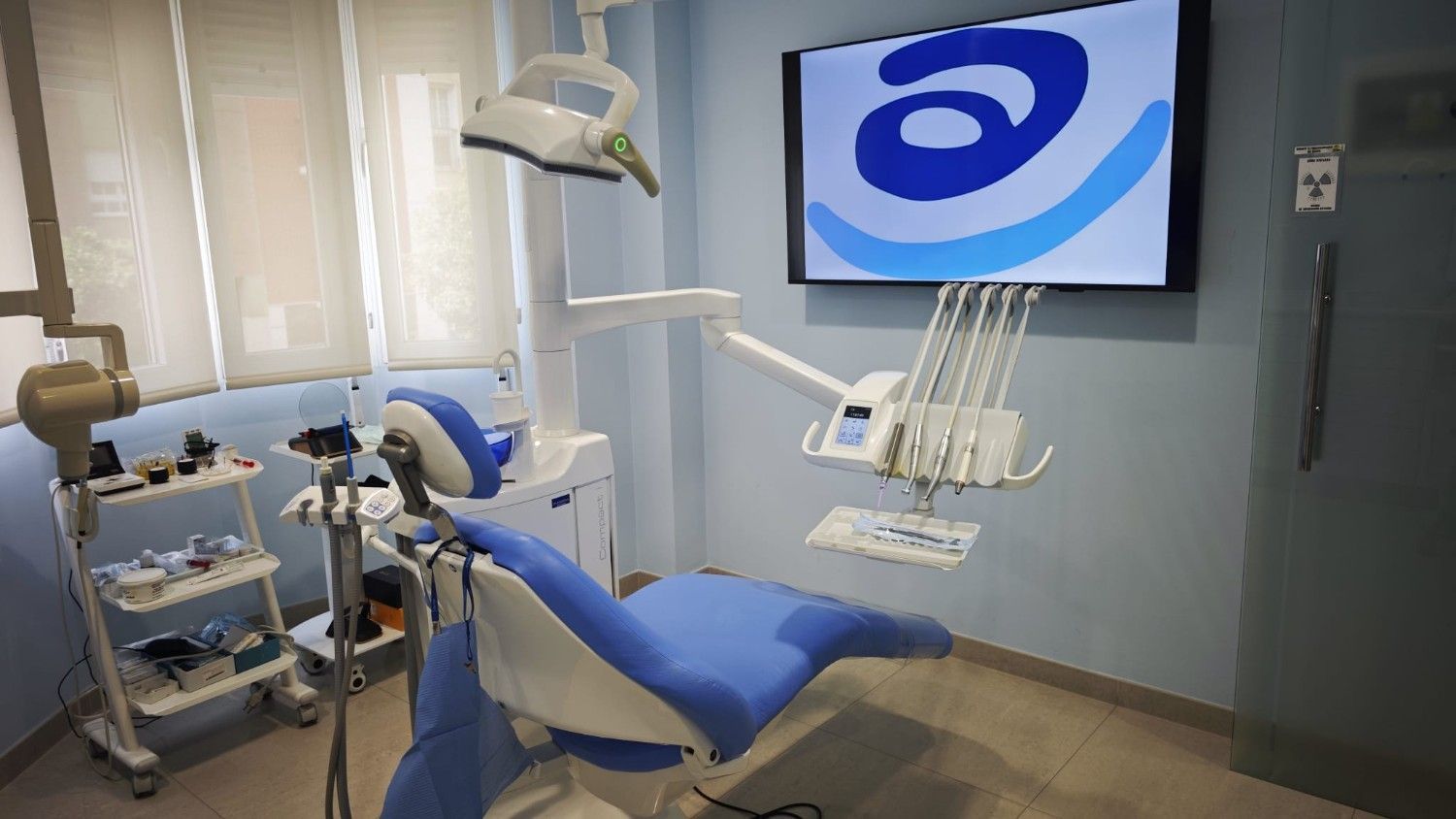Consultorio dental moderno con sillón azul, instrumental y monitor montado en la pared que muestra un logotipo.