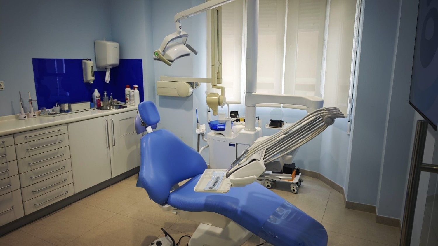 Consultorio dental con sillón dental azul, lámpara de exploración, armarios y persianas en las ventanas.