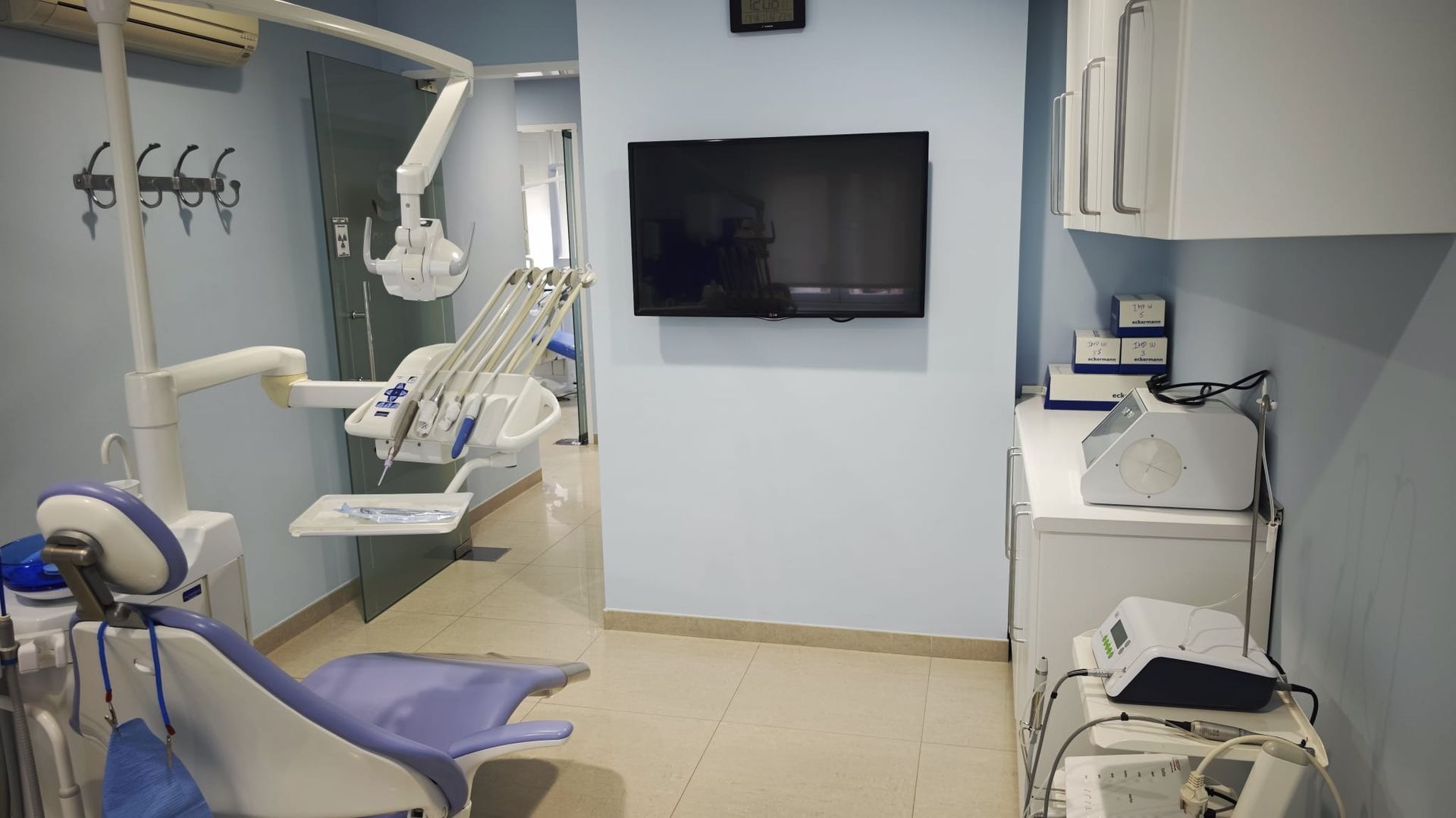Sala de examen dental con sillón, luz de techo, televisor montado en la pared y área de lavabo.