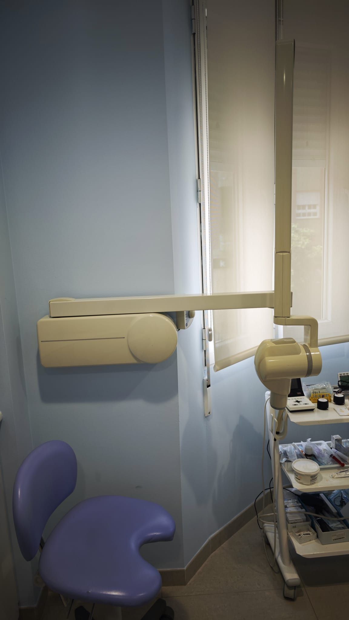 Sala de examen dental con silla azul, brazo de rayos X beige y brazo para equipo montado en la pared.