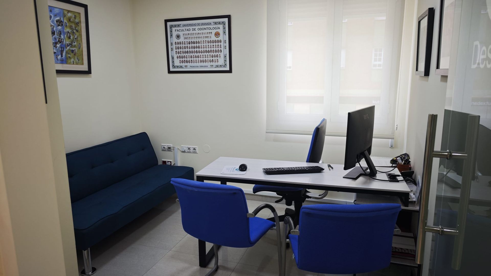 Pequeña oficina con sillas azules, escritorio, monitores de ordenador, sofá y cuadros en la pared.