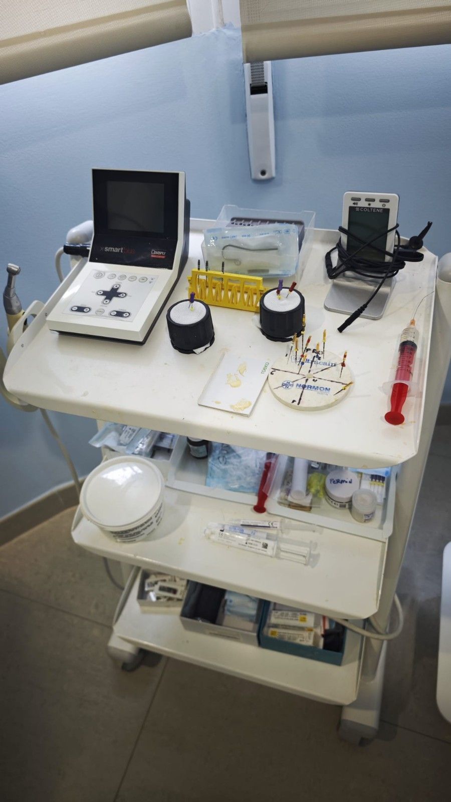 Equipo médico en un carrito con ruedas, que incluye un monitor, cables, contenedores e instrumental, en una sala clínica.