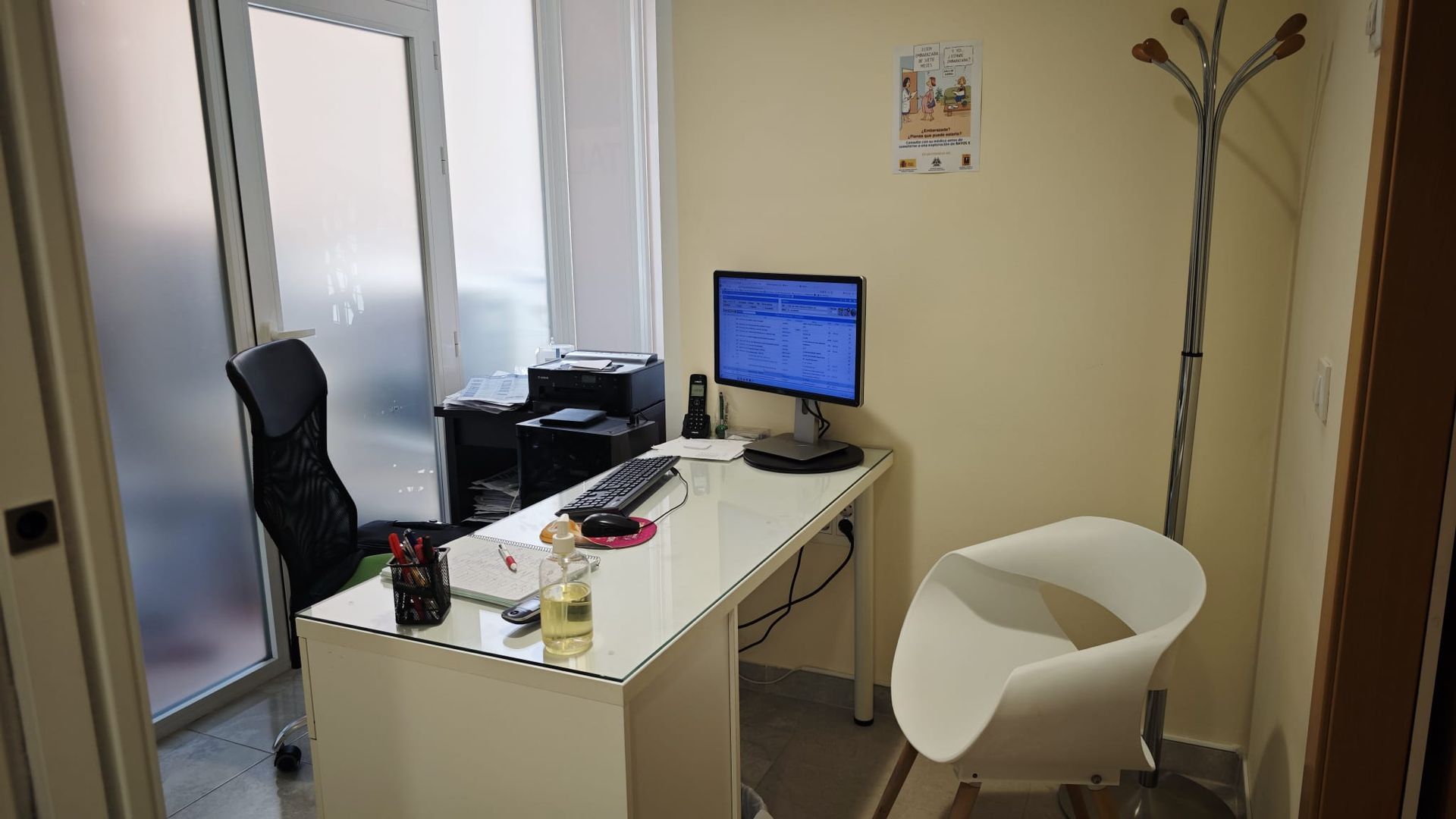 Pequeña oficina con escritorio, monitor de ordenador, silla negra, silla blanca y perchero junto a la pared.